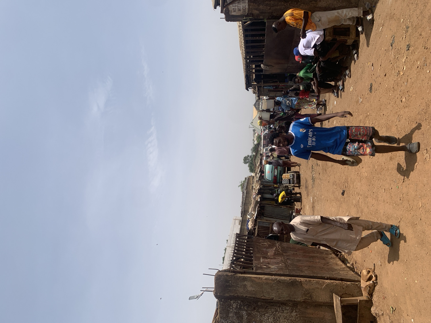 The image depicts an outdoor scene in what appears to be a bustling market or street in Rafin Par, Nigeria, under a bright, clear sky. The ground is unpaved, consisting of reddish-brown dirt.
In the foreground, slightly to the left, a man wearing a light-colored, long-sleeved traditional tunic and light blue shoes stands near a low concrete wall or fence. Further back, towards the center, a young man in a blue "Emirates Fly Better" football jersey (featuring the Real Madrid crest and Adidas logo) and colorful patterned shorts stands facing forward, with his arms slightly out.
The background is filled with numerous makeshift stalls or small shops constructed from corrugated metal, wood, and other basic materials, suggesting a commercial area. Several other people are visible within and around these structures, some sitting on benches, others standing. A light-colored vehicle, possibly a small car or tricycle, can be seen among the stalls.
The bright, pale blue sky, with only a few faint wisps of clouds, indicates a sunny daytime setting, likely mid-morning or early afternoon. The scene captures a moment of daily life, with people gathered, possibly engaging in trade or social interaction. On a small sign attached to a structure in the upper right background, fragmented text is visible, including "NO F...", "WET FO...", and "RACE W...".