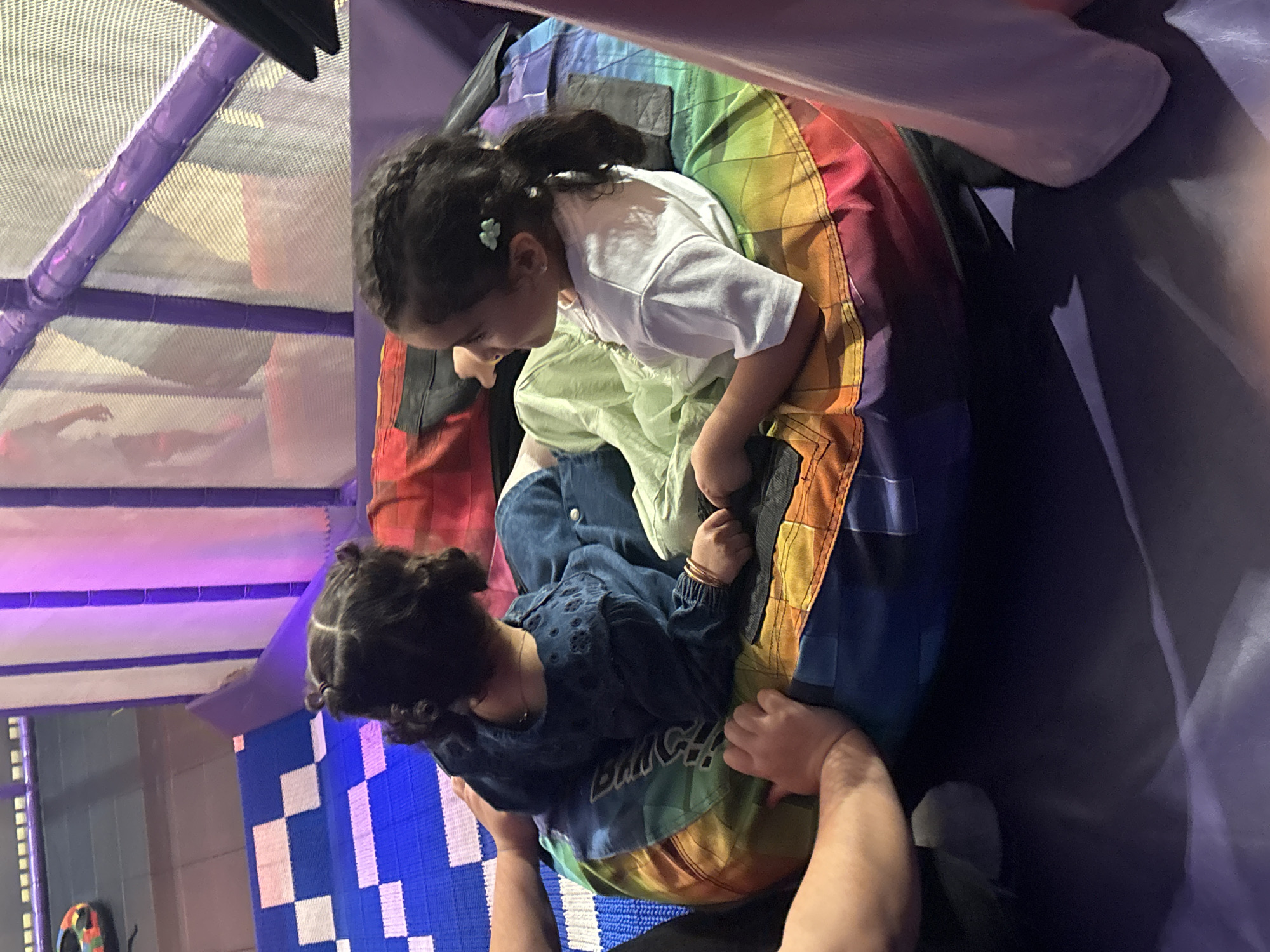 The image shows two young girls sitting on a large, rainbow-colored inflatable cushion in an indoor play area. The girl on the left is wearing a dark blue denim dress and has her dark hair styled in two pigtails. The girl on the right is wearing a light green outfit and a white t-shirt, with her dark hair in a ponytail adorned with a light green flower clip. Both girls are facing each other and appear to be interacting. In the background, there are purple bars and netting, suggesting a climbing or play structure. To the left of the cushion, a blue and white checkered mat is visible, and further back, a colorful toy car can be seen. The overall environment suggests a children's entertainment center. The scene is likely located in Dubai, United Arab Emirates, given the context provided.