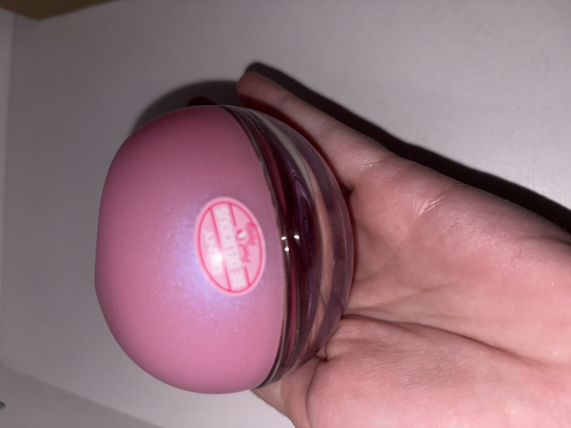 A pink, spherical perfume bottle labeled "DKNY BE DELICIOUS" is held in a person's palm. The bottle has a clear glass base and a pink, pearlescent cap. The background is a plain, light-colored surface. This image was taken in the city of Zasеchnoye, Russia.