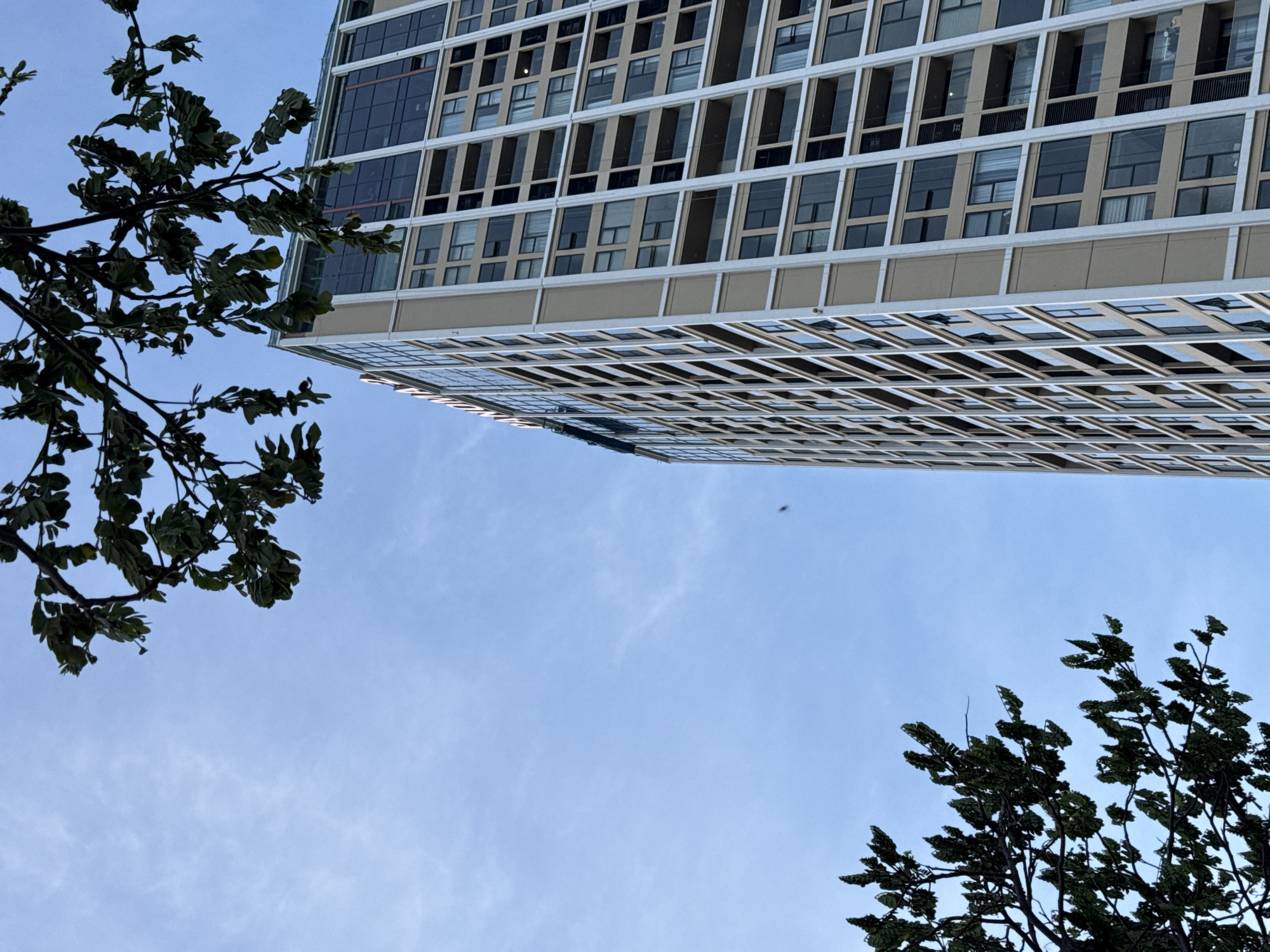 The image shows a tall, modern residential building in Da Nang, Vietnam, with a clear blue sky in the background. The building features a beige facade with a grid of windows and balconies. There are trees with green leaves in the foreground, partially obscuring the view of the building and the sky. The angle of the shot is from below, looking up towards the building and the sky. There are no people visible in the image.
