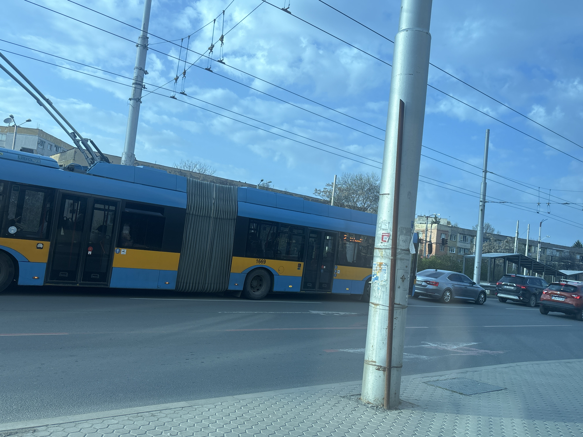 The image shows a street scene in Sofia, Bulgaria. A blue and yellow trolleybus, numbered 1669, is traveling along the road. Behind it, a silver Skoda car with license plate CB 9294 BA, a black Mitsubishi Outlander car with license plate CB 4981 BX, and a red car with license plate CB 6934 BB are traveling. A grey metal pole with advertisements affixed is in the foreground. Overhead are numerous electrical lines and support structures for the trolleybus system. Buildings are visible in the background, including what appears to be apartment blocks. The sky is blue with scattered clouds. A bus stop shelter is seen further down the road. The photograph was taken from the sidewalk.