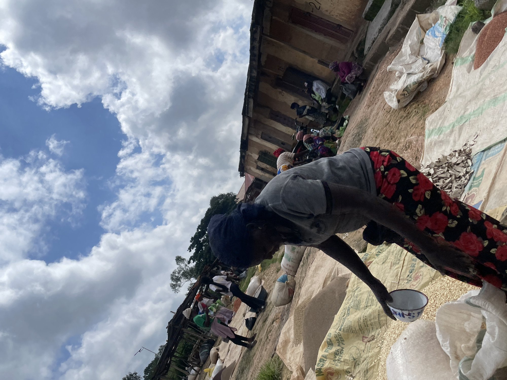 The image depicts an outdoor market scene in the city of Wang, Nigeria, under a cloudy sky. In the foreground, a woman wearing a grey t-shirt and a black skirt with a red floral pattern is pouring something from a white and blue bowl into a large sack of grain. She is positioned on a slope covered with various grains and sacks. To the left and further up the slope, other people are visible, some interacting with sacks of grain. In the background, there are structures that appear to be market stalls or buildings, with wooden roofs and walls. The ground is predominantly dirt and covered with scattered materials, including sacks and piles of grain. Several people are seated or standing around the buildings, some appearing to be vendors or customers. The overall impression is a bustling, informal marketplace where agricultural goods are being traded.