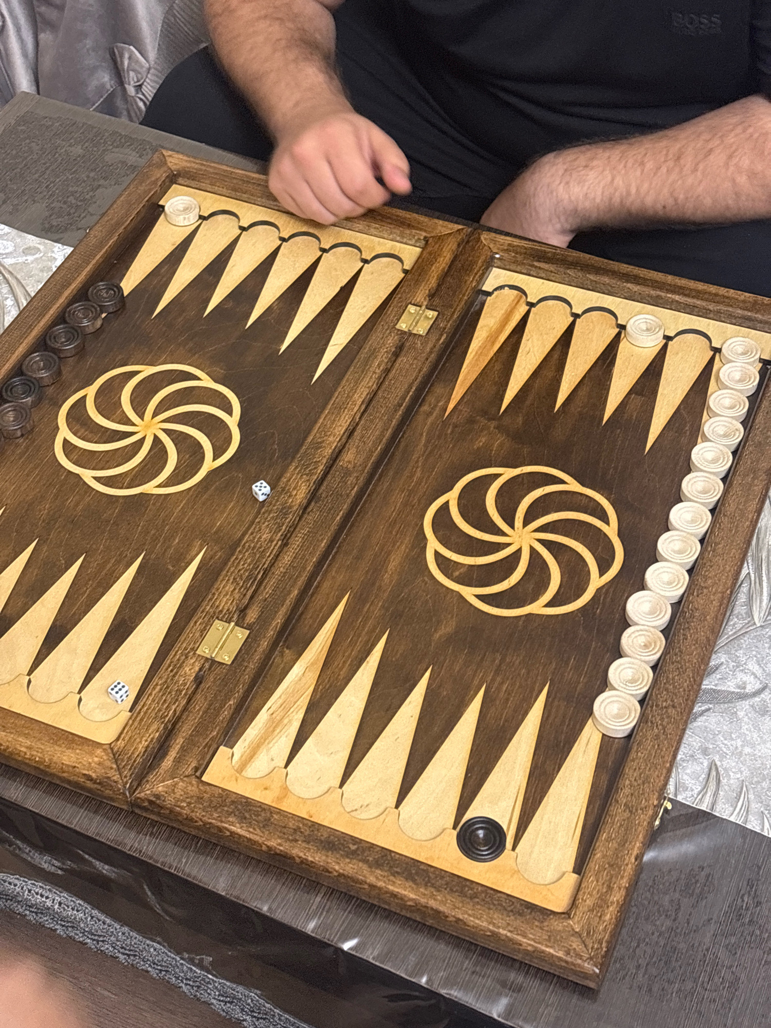 The image shows a close-up, overhead view of a wooden backgammon board, open and lying on a table. The board is dark brown with light wood triangular points. There are two sets of playing pieces: dark brown and light cream-colored checkers. Two dice are visible on the board. A person's hands and arms are partially visible, suggesting they are playing the game. The background is blurred but appears to be an indoor setting with furniture. The scene is set in the city of Getahovit, Armenia.