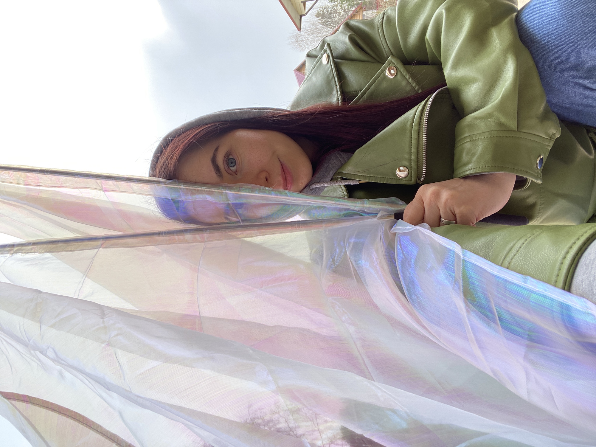 The image captures a close-up, low-angle perspective of a young woman lying down or leaning back, looking directly at the viewer with a gentle, inviting smile. She has long, dark reddish-brown hair, blue eyes, and visible freckles on her nose and cheeks. She is wearing a sage green leather-like jacket with metallic snap buttons and a zipper, over a grey hooded sweatshirt. Part of her blue jeans is also visible. A silver ring adorns a finger on her visible hand, which appears to be holding onto something or resting on the iridescent fabric.
A translucent, iridescent fabric, possibly organza or sheer material, dominates the foreground and midground, draped and folded around the woman. This fabric reflects pastel hues of pink, purple, blue, and green, creating a shimmering, ethereal effect. It appears to be suspended from a thin rod or frame, creating a soft, diffused barrier between the camera and the subject.
The background shows a bright, overcast sky. Hints of building structures with reddish roofs and bare tree branches are visible, suggesting an outdoor setting, likely in a residential or suburban area. The soft, diffuse lighting indicates daytime, possibly on an overcast day. The overall mood is calm and serene, with the woman's expression conveying warmth and approachability. The location context for the image is Фоки, Russia. No visible text is present in the image.