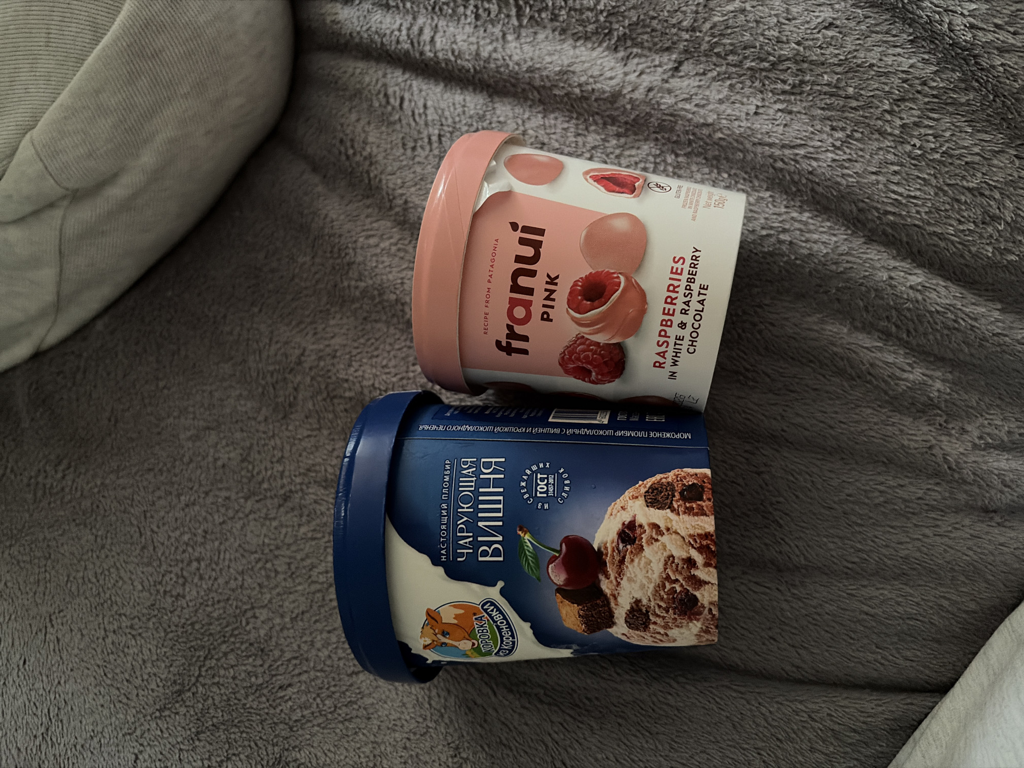 Two containers of frozen treats are positioned side-by-side on a textured, dark gray blanket, possibly a bedspread or throw, in a location identified as the city of Новинки, Russia. The container on the left is predominantly blue and white, labeled "КОРОВКА из Кореновки" (KOROVKA from Korenovka) and features the flavor "ЧАРУЮЩАЯ ВИШНЯ" (CHARMING CHERRY) with an image of cherry-swirled ice cream, a fresh cherry, and chocolate brownie pieces. Text indicates it is "НАСТОЯЩИЙ ПЛОМБИР" (REAL PLOMBIR) and adheres to "ГОСТ 31457-2012". The container on the right is pink and white, branded "franui PINK," and advertises "RASPBERRIES IN WHITE & RASPBERRY CHOCOLATE," displaying images of whole raspberries and raspberry-filled chocolates. It states "RECIPE FROM PATAGONIA" and "GLUTEN-FREE," with a net weight of 150g. A faint, lighter fabric is visible in the upper right and lower left background, suggesting additional soft furnishings.