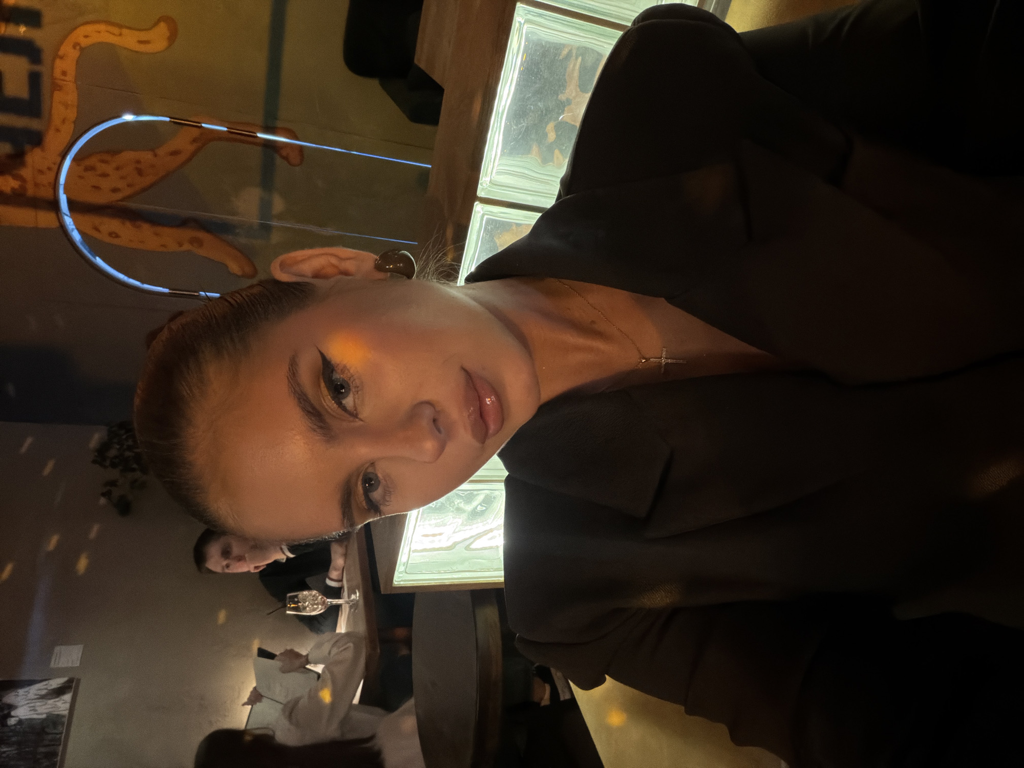 A selfie of a woman with blonde hair and fair skin. She is wearing a black blazer and a gold necklace with a small cross pendant. The background features a wall with a decorative light fixture resembling a leopard, a wall made of glass blocks, and a dimly lit restaurant setting with other patrons visible. A man can be seen in the background, sitting at a table with a glass of wine. The image was likely taken indoors at a restaurant or bar in Krasnoyarsk, Russia.