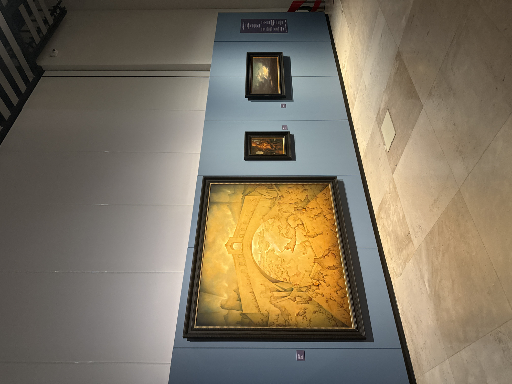 The image shows an exhibit of paintings in a museum setting. Three framed paintings are vertically arranged on a pale blue wall. The largest painting at the bottom depicts a golden-toned scene with a bridge or arch-like structure dominating the composition. The upper paintings are smaller, with the uppermost one featuring a dark landscape or seascape and the middle one containing abstract or indistinct figures. Above the top painting is a panel with written information, presumably about the artworks or the artist. The setting is a modern building with clean lines, with a white or light grey wall on the left and a light-colored, tiled floor or wall on the right. The location of the city of San Bernardino Tlaxcalancingo, Mexico is not shown. There are no people in the image.