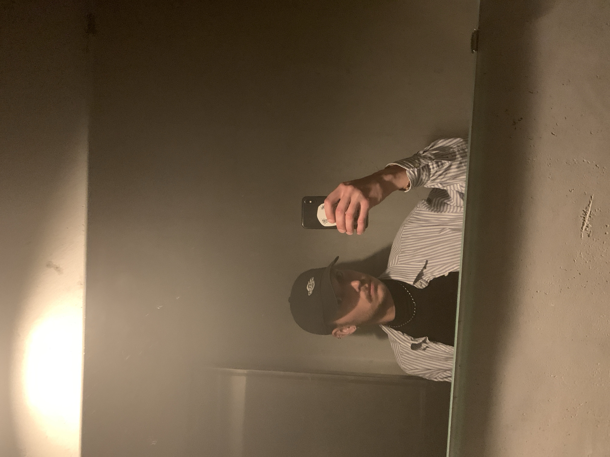 A person takes a mirror selfie in a bathroom with gray walls. The person wears a black baseball cap, a black shirt, a striped collared shirt, and a silver chain. The person is holding a phone. The location is unidentifiable as Tel Aviv, Israel.
