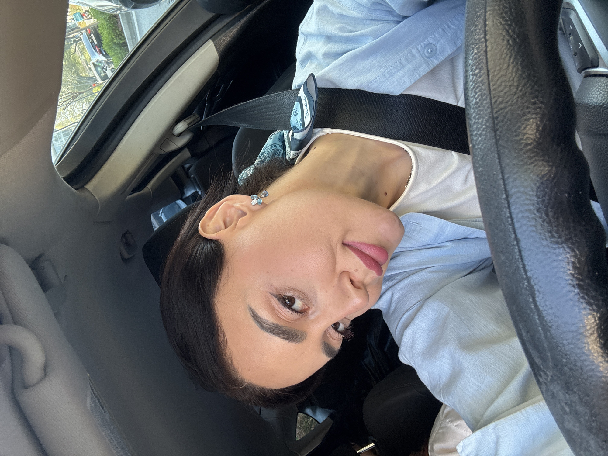 A medium shot shows a woman seated in the driver's seat of a car during daylight hours. The image is presented sideways, rotated 90 degrees counter-clockwise. The woman, with dark hair and well-defined eyebrows, looks directly at the camera with a calm expression and a hint of a smile, her lips adorned with pink lipstick. She is wearing a light blue button-up shirt layered over a white top, with a black seatbelt fastened across her chest. Attached to the seatbelt is a distinctive fabric accessory, primarily white and blue, featuring the visible text "Bluetable." A light blue and silver floral earring is visible on her ear, and two small moles are noticeable on her upper chest/neck area.
The black steering wheel is visible on the right side of the frame. Through the car's windows, a bright, sunny outdoor scene unfolds, revealing trees lining a street and a silver/grey car in the distance, indicative of an urban or suburban environment. The car's interior appears dark, contrasting with the bright light from outside. The setting is in Tbilisi, Georgia.