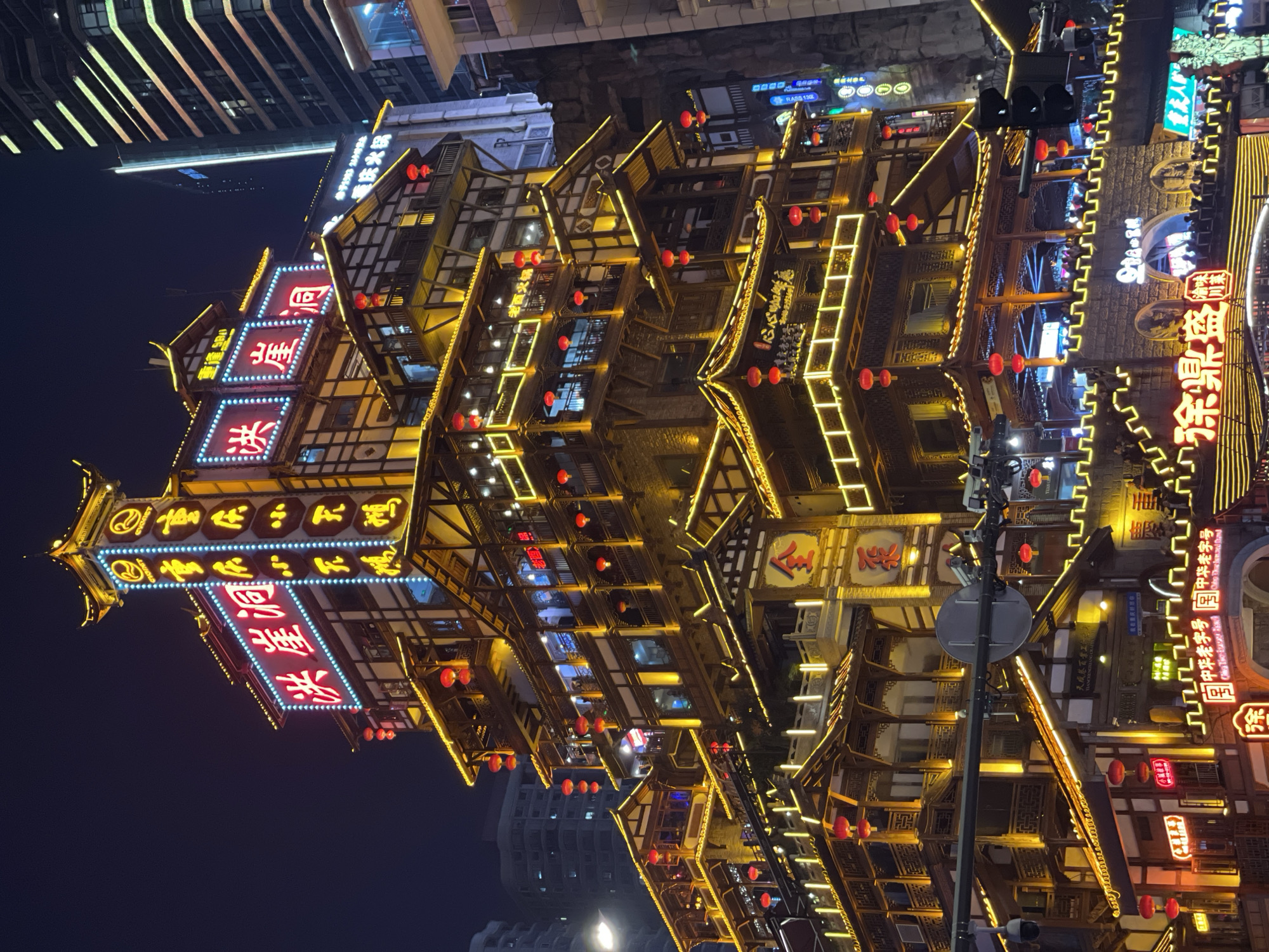 The image depicts the Hongyadong commercial complex at night, located in Yuzhong District, Chongqing, China. The scene is dominated by a multi-tiered, stilt-house style building, illuminated extensively with golden LED strip lights outlining its traditional wooden architecture, numerous red spherical lanterns, and prominent red neon signage.
The primary structure is characterized by its traditional Chinese architectural elements, including multiple curved roofs, intricate wooden balconies, and a facade featuring white plaster walls contrasting with dark brown timber frames. Large red neon signs bearing the characters "洪崖洞" are prominently displayed on multiple levels, with additional illuminated signage such as "重慶" and "老重慶火鍋." A tall, vertical sign tower on the upper right of the complex displays blue and yellow illuminated text and a "CYGNET" logo.
Multiple open-air balconies and terraces are populated with numerous individuals, suggesting a bustling public space or entertainment venue. People are visible standing and observing, indicating social activity.
In the foreground, a stone bridge-like structure with crenelated walls and archways is also illuminated with golden lights, with more people visible atop it. Street furniture includes a black, vertically oriented traffic light system on the right and a circular road sign in the lower-middle, partially obscured by overhead lines. Security cameras are mounted on poles.
In the background and to the right, modern high-rise glass and steel skyscrapers are visible, some with illuminated windows and vertical light strips, providing a stark architectural contrast to the traditional complex. The sky is dark, indicating a nighttime setting.