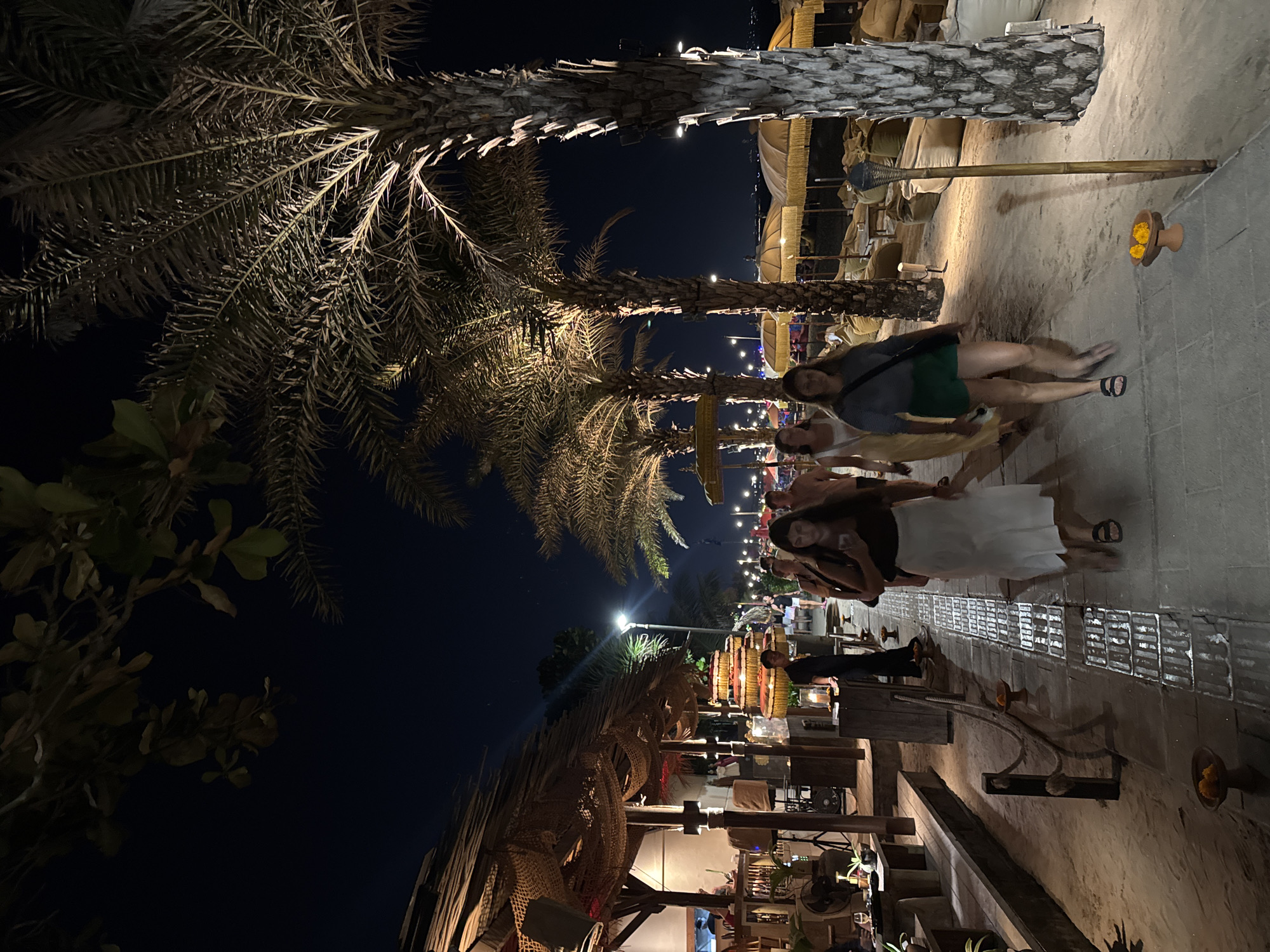 This is a night-time, eye-level, medium shot of a beachside walkway in Kuta, Indonesia. In the foreground, three women are walking away from the camera along a paved path illuminated by artificial lighting. The woman on the left wears a white skirt and a dark top, the middle woman wears a yellow dress, and the woman on the right wears green shorts and a dark top. Palm trees line the right side of the walkway, and to the left, a thatched-roof structure with seating and lighting is visible. In the background, beach umbrellas and lounge seating can be seen, suggesting a resort or restaurant setting. The sky is dark, indicating it is nighttime. The overall scene is a bustling outdoor dining or recreational area at night.