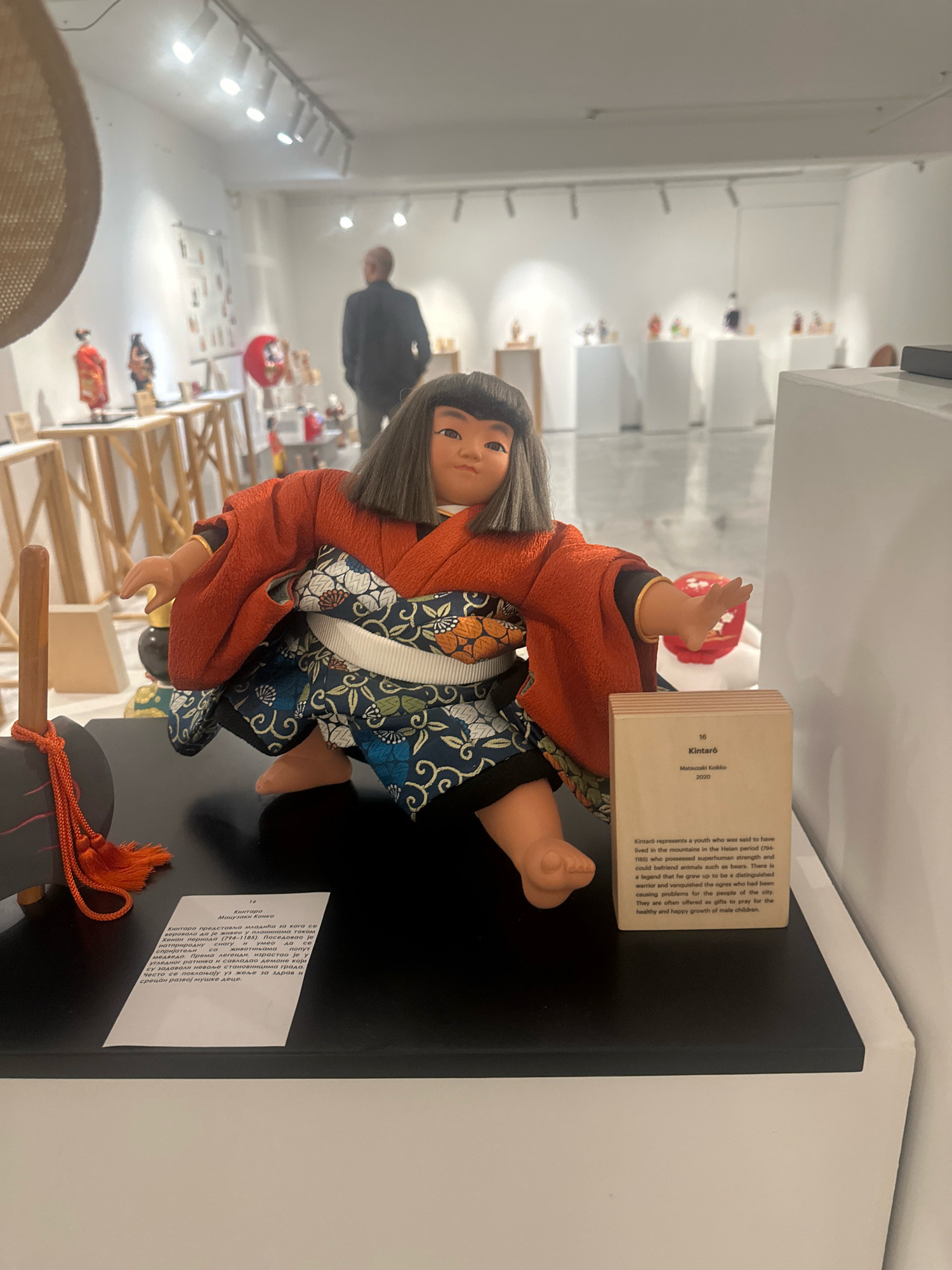 In the foreground of the image, a Japanese doll is displayed on a black surface. The doll is dressed in a traditional orange kimono with blue, white, and green floral patterns on its underskirt. It has dark, bobbed hair and a round face with painted features. To the right of the doll is a small, beige booklet with text on it, and to the left, a dark object with an orange tassel. In the background, the scene appears to be an art gallery or exhibition space. White walls are visible, along with track lighting on the ceiling. Several other displays with artwork and smaller objects are set up on white pedestals and wooden tables. A man in a dark jacket is standing in the middle ground, facing away from the camera, looking towards the exhibits. The overall environment suggests a public exhibition, possibly of dolls or cultural artifacts.