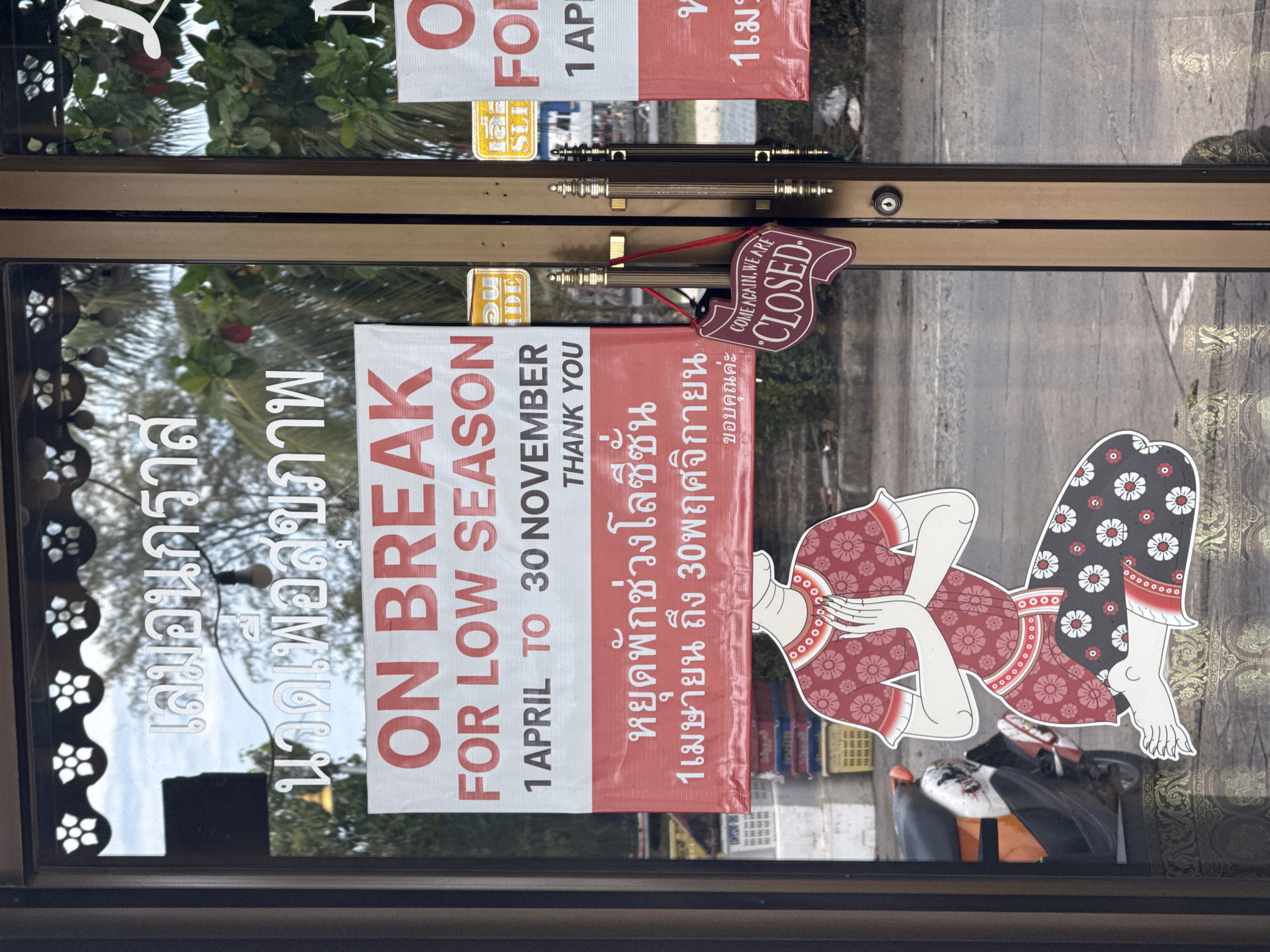 A shop in Thong Sala, Thailand, is closed for the low season, as indicated by a sign on the door written in both English and Thai. The sign reads "ON BREAK FOR LOW SEASON 1 APRIL TO 30 NOVEMBER THANK YOU" in English. Below this, the same message is written in Thai. The door also has a "COME AGAIN, WE ARE CLOSED" sign. A sticker of a traditional Thai dancer is affixed to the door, along with the shop name in Thai above the signs. The shop's dark brown door frame is visible.