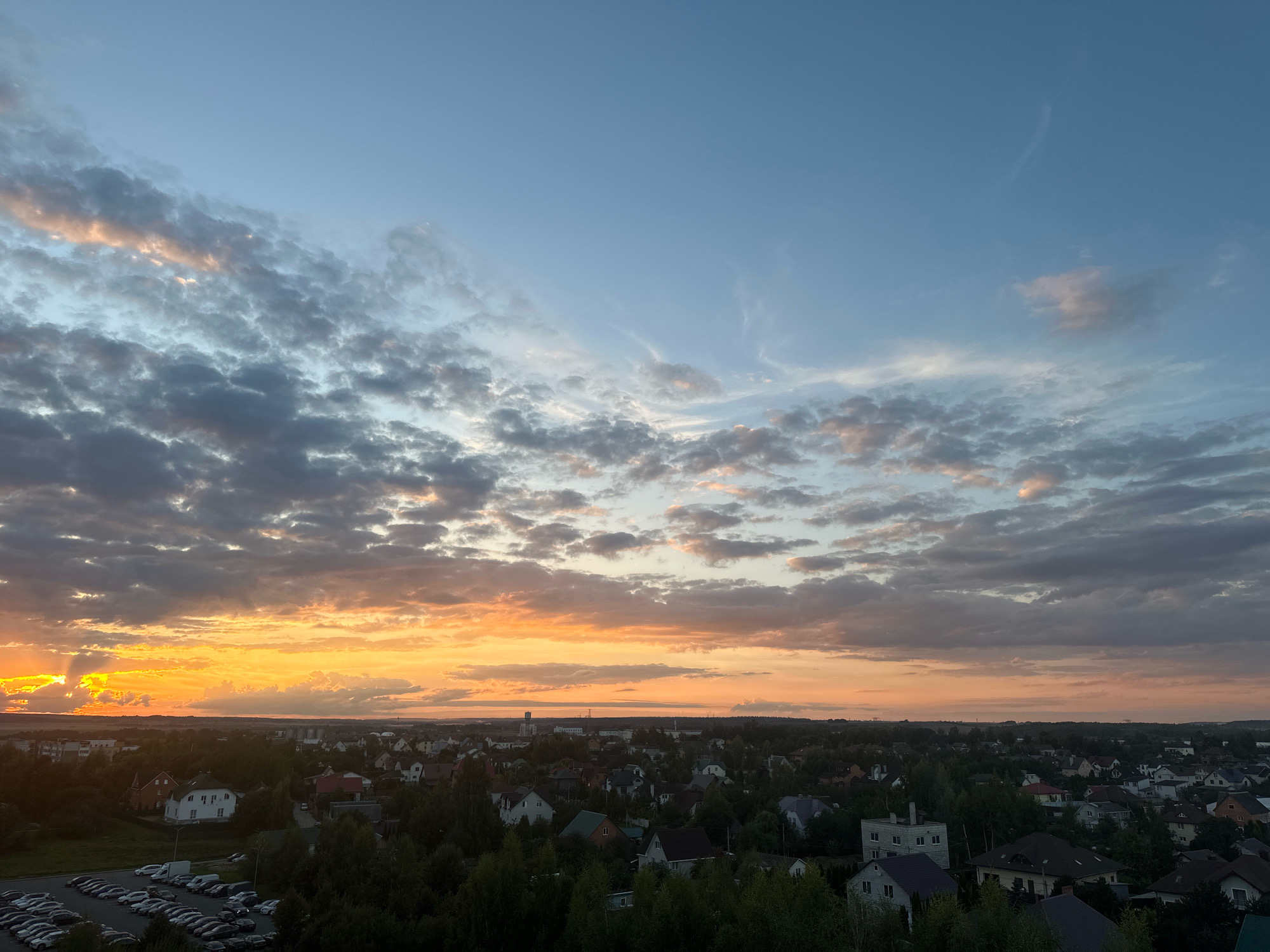 The image presents an aerial view of a residential area at sunset, likely within the Шчомысліцкі сельскі Савет in Belarus. The sky is a dramatic canvas of orange and yellow hues from the setting sun, contrasting with the blue expanse above. Wispy clouds are scattered across the sky, with some catching the golden light. Below, a dense collection of houses with varying roof colors and architectural styles fills the landscape, interspersed with lush green trees. A parking lot filled with numerous cars is visible in the foreground, on the left side of the image. A water tower or similar tall structure is present in the distance, and the horizon suggests a flat terrain extending beyond the immediate settlement. The overall scene depicts a tranquil evening in a suburban or rural setting.