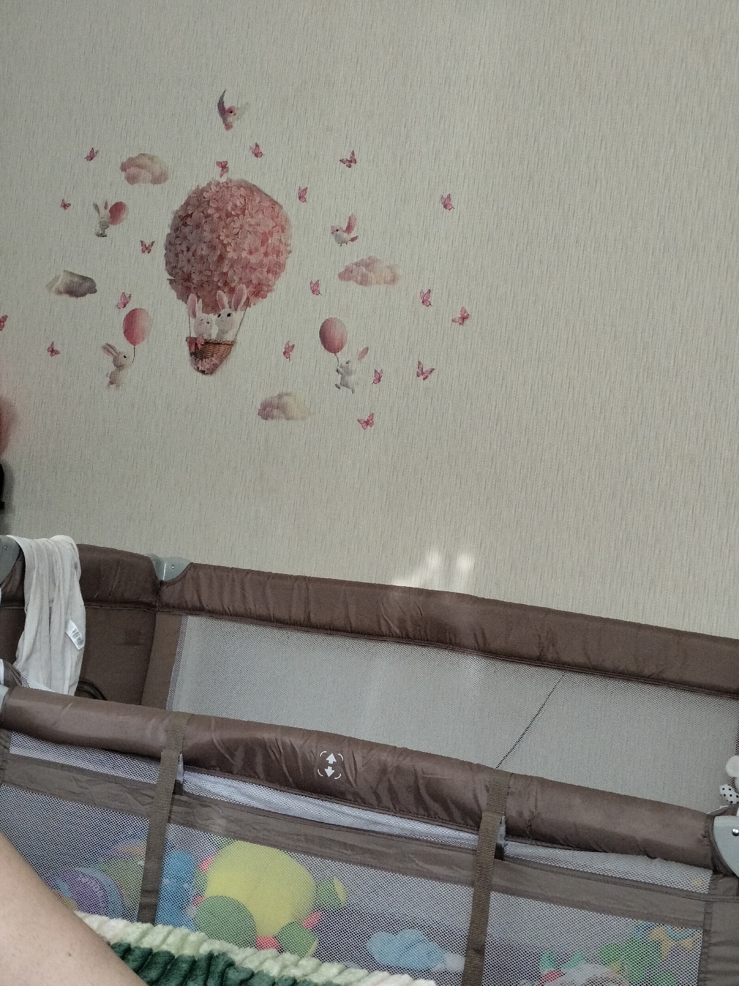 The image shows a close-up view of a baby crib with decorations on the wall behind it. The wall has a textured, light-colored wallpaper with a pattern of faint vertical lines. Adorning the wall are several decals depicting a whimsical scene. A large, round, pink hot air balloon, made of flower-like shapes, is the central element. Two white bunnies with long ears are in the basket of the hot air balloon. Other decals include smaller pink balloons, pink butterflies, and fluffy pink clouds. A few scattered bunnies are also depicted, some holding balloons and appearing to be floating or flying. The overall theme of the wall decorations is playful and childlike.
The crib itself is brown with a mesh netting on the sides, allowing a view of colorful toys inside, including a yellow ball and what appear to be other soft toys in blues and greens. A white cloth is draped over the side of the crib. A strap from the crib is visible in the center of the image. The lighting suggests it is daytime, possibly indoors with natural light filtering in, casting some shadows and reflections on the crib's mesh. The scene evokes a nursery or child's room, likely in a home setting in Vysokyi, Ukraine. There is no visible text in the image.
