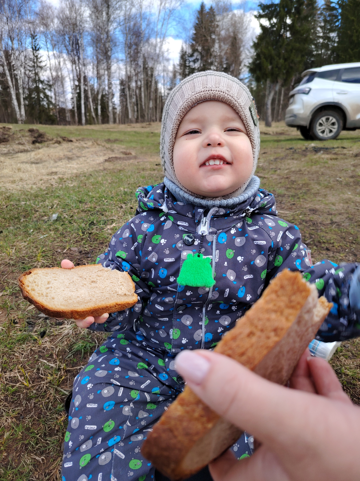 The image shows a young child sitting on the grass, holding a piece of bread in their left hand and another piece being offered to them by a person out of frame. The child is wearing a grey and beige knit hat and a blue and grey patterned snowsuit. They are smiling slightly, showing their teeth. The background consists of a grassy field, trees, and a white SUV parked in the distance. The environment suggests an outdoor setting, possibly a park or rural area.