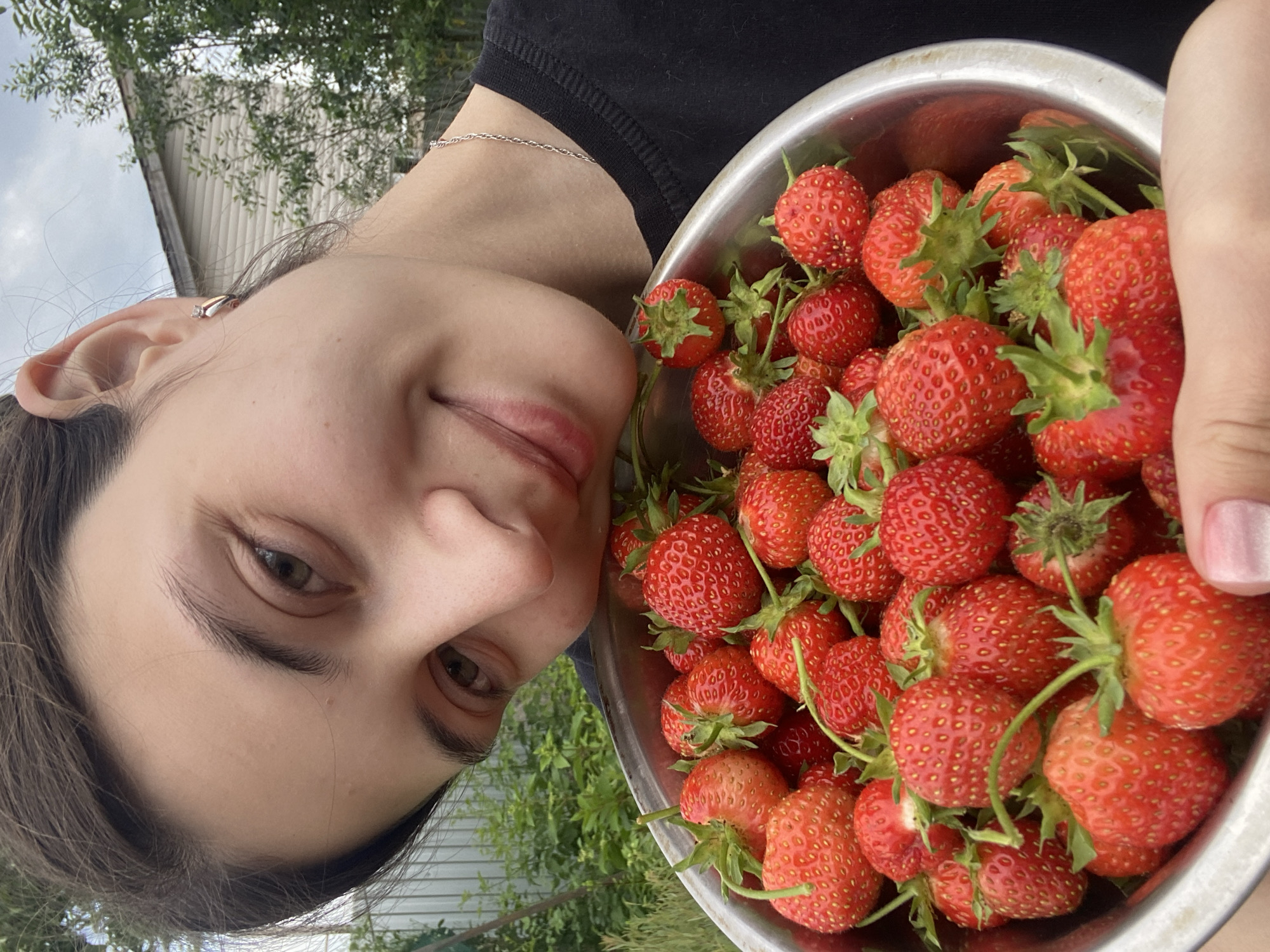 The image is a close-up, low-angle selfie of a young person holding a metal bowl overflowing with fresh strawberries. The person's face is partially visible, tilted towards the camera with a gentle smile. They are wearing a black t-shirt and a thin silver chain around their neck. A small, sparkling earring is visible on their left ear. The strawberries are a vibrant red, with green stems and leaves, filling the bowl and spilling over the sides. The background shows greenery, suggesting an outdoor setting, possibly a garden or yard, with hints of a building and a cloudy sky, indicating it's likely daytime. The overall mood is cheerful and summery. No text is visible in the image. The metadata indicates the location context is Votkinsk, Russia.