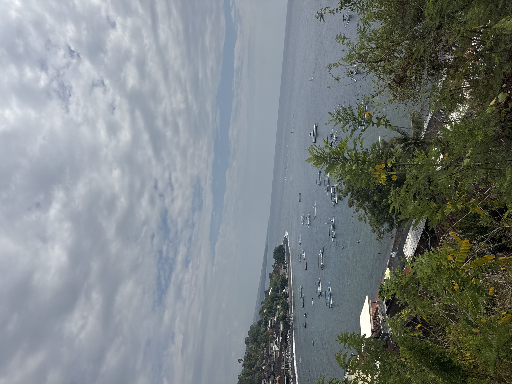 The image shows a coastal view of Abang, Indonesia, with a village situated on a hillside overlooking a bay filled with numerous small boats. The sky is overcast with scattered clouds, and the sea is a dark blue. The village appears to have traditional Balinese architecture with terracotta roofs. In the foreground, lush green foliage obscures some of the view. Several people can be seen in the water near the shore, and some are on the beach. The boats are mostly traditional outrigger canoes, commonly used in Indonesian waters. The overall scene is tranquil and depicts a typical day in a coastal Indonesian village.
