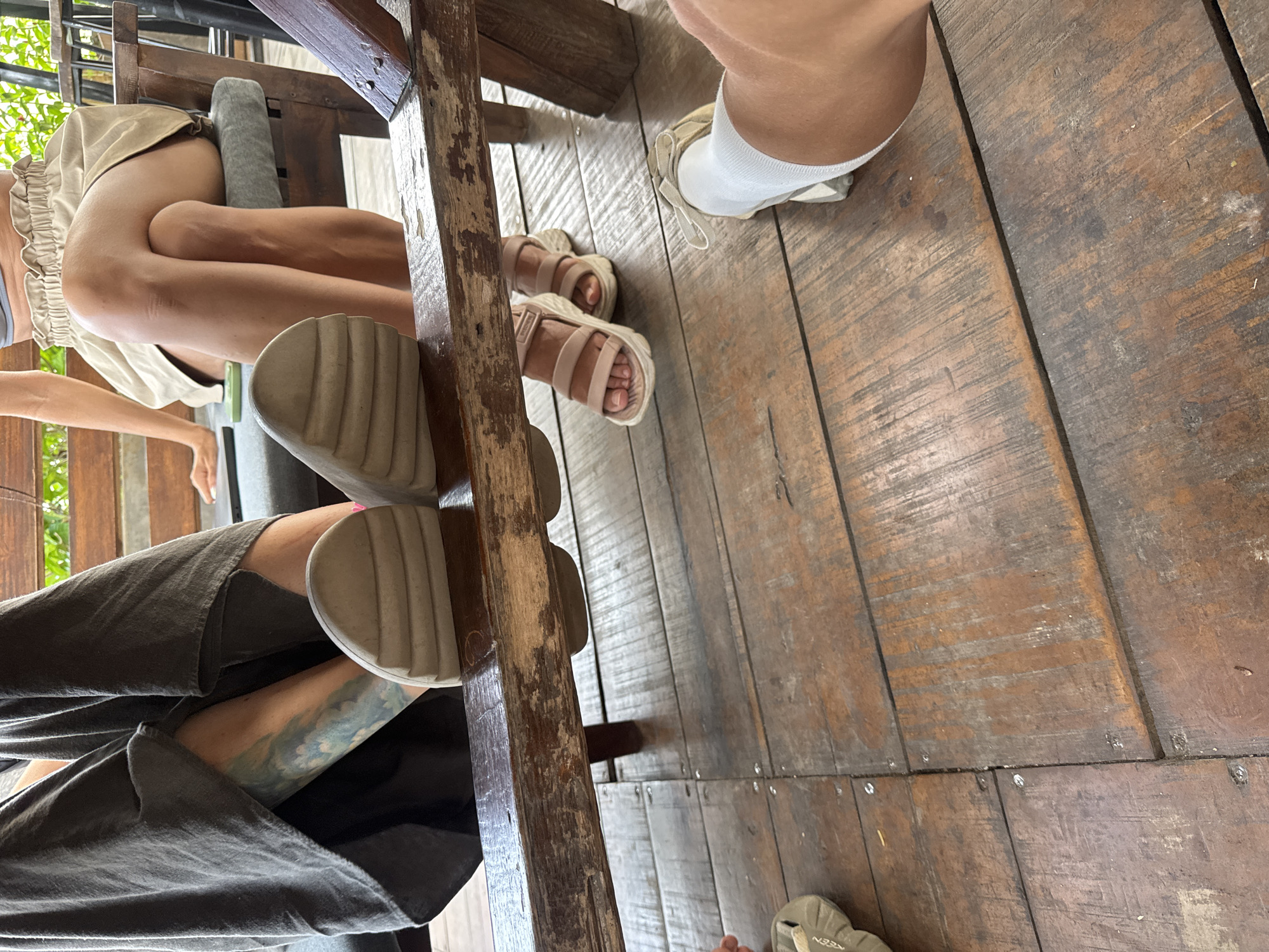 The image presents a ground-level view of several people's legs and feet in a rustic, casual setting, likely in Hà Giang 1 Ward, Vietnam. The main subjects are at least three individuals seated around what appears to be a sturdy wooden table or bench, indicated by the prominent dark wooden beam running through the center of the frame and other wooden structures.
One person, positioned towards the top left, wears light-colored shorts with a ruffled hem and light beige, chunky-soled sandals. Their bare legs are visible. Another individual, in the bottom left, wears dark, loose-fitting trousers that partially cover a distinctive blue and green tattoo on their lower leg. They are wearing light-colored, thick-soled sandals, one of which rests against a wooden support. A third person's feet are visible on the right side of the image, wearing light-colored socks and an off-white shoe or sandal, with "KEEN" faintly visible on the sole. A fourth person's arm and hand are briefly seen on the far left, reaching towards the floor.
The setting features worn, dark wooden floorboards with visible grain and small nails or screws. The overall lighting suggests it is daytime. Green foliage is visible in the background on the far left, indicating an outdoor or semi-outdoor environment, possibly a café, restaurant, or home with an open-air area. The activity appears to be a relaxed social gathering or a meal, with people seated comfortably. The rough-hewn wooden elements contribute to a natural and unpretentious ambiance.