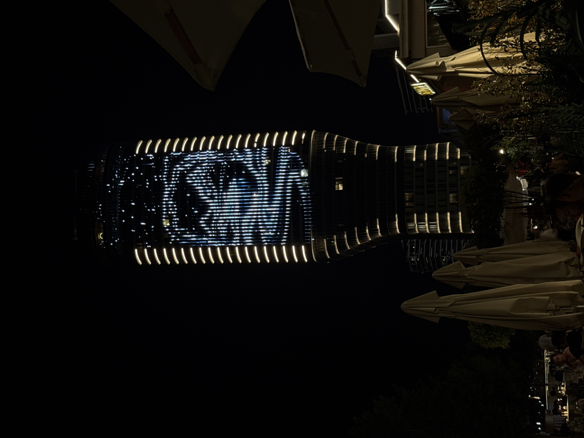 The image captures a nighttime scene in Belgrade, Serbia, dominated by a tall, modern skyscraper illuminated with a dynamic light display that resembles a pixelated face. The building, likely part of the Belgrade Waterfront development, features vertical lines of warm white lights along its edges and a central facade covered in a pattern of blue and white lights. In the foreground, several closed outdoor umbrellas, a common sight at restaurants and cafes, are clustered together, their light beige fabric contrasting with the dark night. Beyond the umbrellas, patrons are seated at tables in a dimly lit outdoor seating area, suggesting a restaurant or bar. Trees and foliage are visible, adding to the urban oasis atmosphere. The overall impression is one of a vibrant, contemporary urban setting, where architecture and entertainment converge.