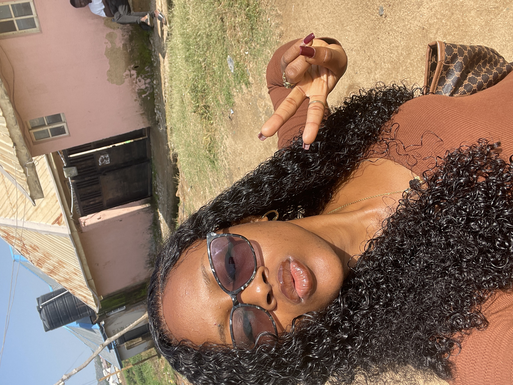 A woman is taking a selfie outdoors in Rafin Par, Nigeria. She is wearing a brown top and sunglasses. Her hair is long and curly, and she has on long, dark red acrylic nails. She is holding up two fingers in a peace sign. In the background, there are houses, some with corrugated metal roofs, and a person sitting on steps outside one of the buildings. The ground is dusty and covered with some dry grass.