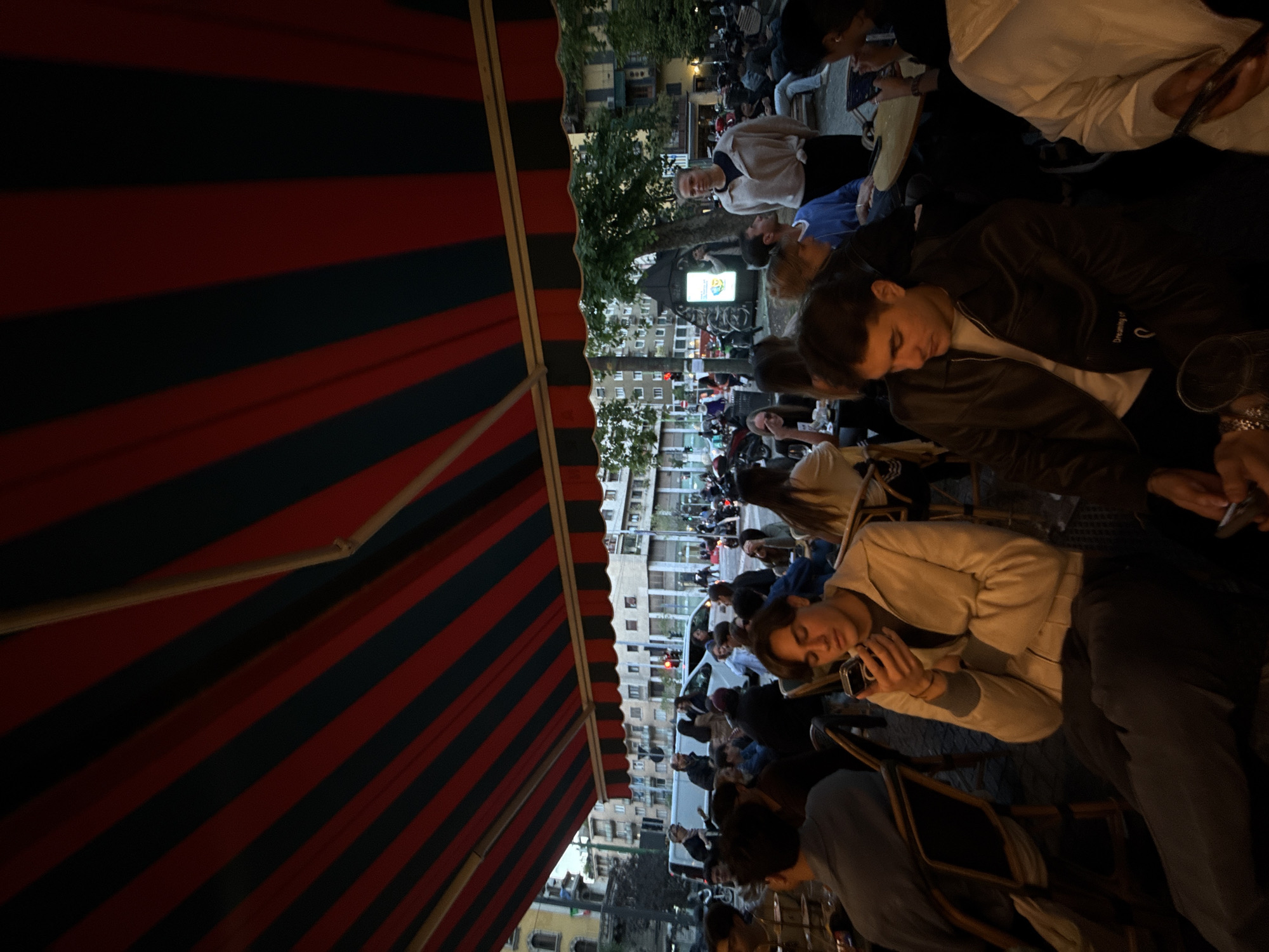 The scene depicts an outdoor gathering, likely at a cafe or restaurant, under a red and dark blue striped awning. People are seated at tables. The setting appears to be a city center with buildings lining the street. Based on the architectural style and ambiance, the location is likely Milan, Italy. People are engaged in conversations and using mobile devices. The overall atmosphere is relaxed and social.