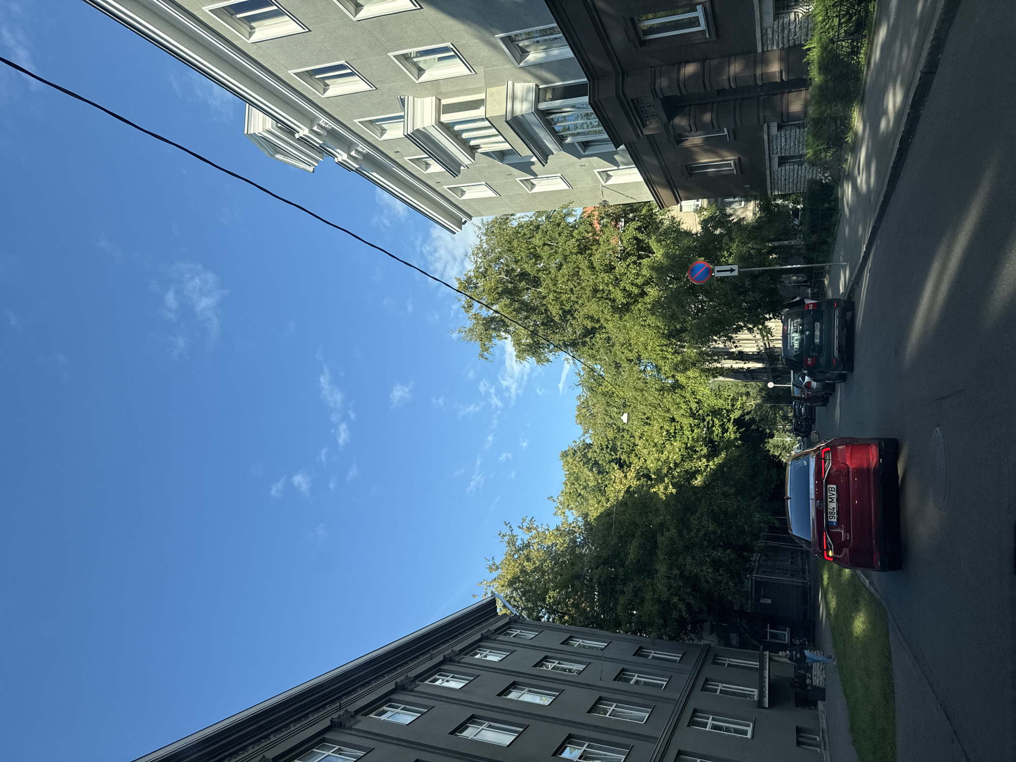 The image depicts a street scene in Tallinn, Estonia, on a clear, sunny day with a bright blue sky and scattered clouds. The perspective is from inside a vehicle, looking forward down a paved road. On the left, a multi-story building with a gray facade and numerous windows lines the street. On the right, another multi-story building with a lighter gray facade, featuring architectural details like a bay window and white trim, is visible. Between the buildings, mature trees with lush green foliage provide shade.
Several vehicles are present on the road and parked along the side. A red sedan with the license plate "984 MVB" is in the foreground, facing away from the camera. Further down the road, a dark-colored SUV is parked on the right. A traffic sign with a red circle and a blue background, indicating a prohibition, is posted on a pole.
A person wearing blue jeans and a light-colored top is walking on the sidewalk in front of the gray building on the left. Shadows from the trees and buildings are cast across the road and sidewalk, indicating the time of day is likely late morning or early afternoon. The overall environment suggests an urban residential or mixed-use area.