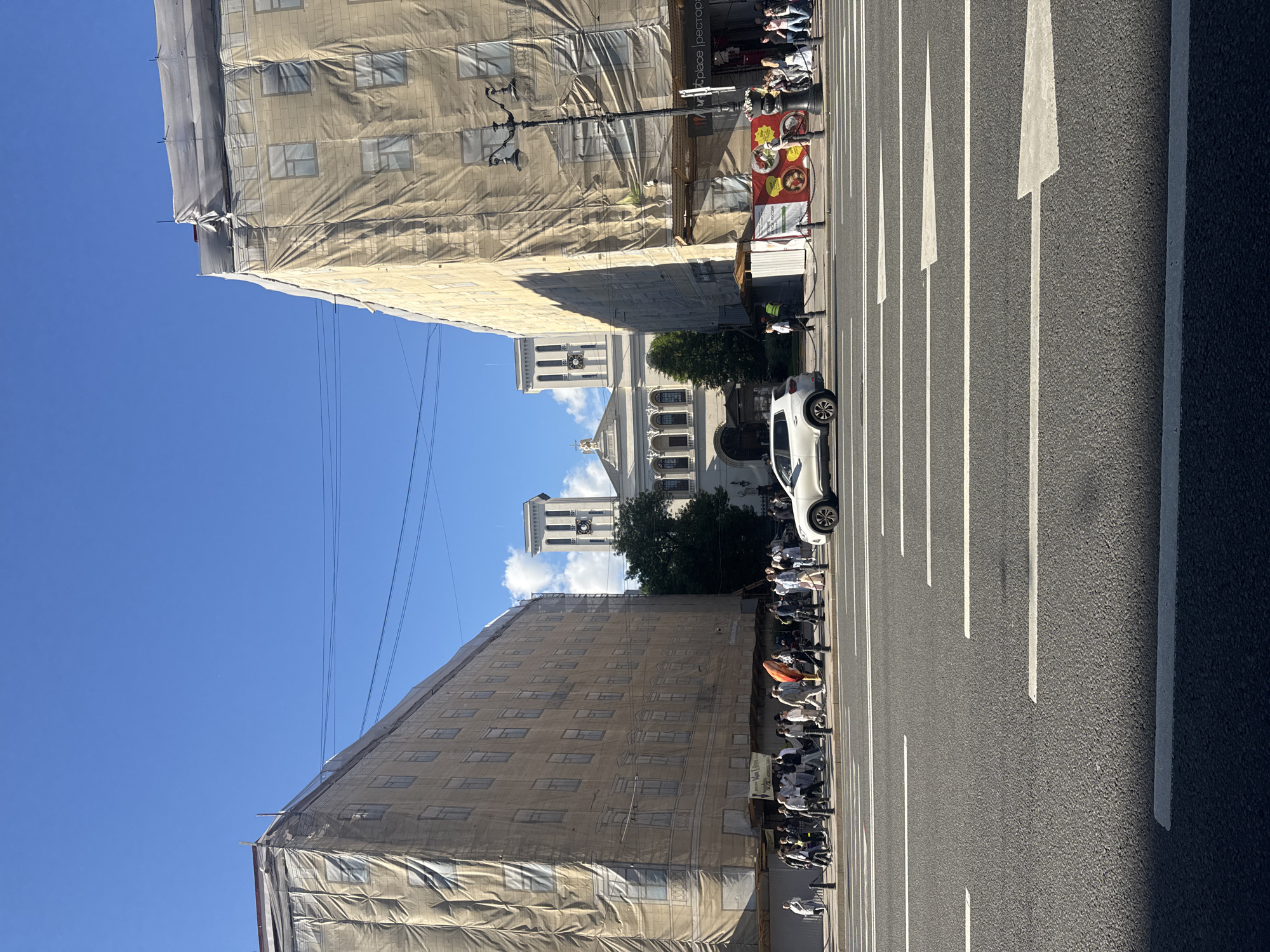 This is a street-level photograph taken in Saint Petersburg, Russia, likely on a sunny day given the clear blue sky with scattered clouds. The perspective is from the middle of a multi-lane asphalt road, with white directional arrows painted on the surface.
On either side of the street, large buildings are covered in tan scaffolding netting, suggesting ongoing construction or renovation. The building on the left is a multi-story structure with visible windows beneath the netting. The building on the right also has a similar scaffolding covering, and a street lamp with decorative elements stands in front of it. A banner with images of food items is visible near the ground level of the right building.
In the distance, between the two draped buildings, a large, ornate white building with classical architecture is visible. It features a central section with arches and columns, topped by two symmetrical square towers with clocks. This building appears to be a significant landmark, possibly a church or a public institution.
A white SUV is driving on the road, and a group of people are walking on the sidewalk in front of the buildings. Some individuals are dressed casually, while others appear to be workers, possibly in high-visibility vests. The overall impression is a busy urban scene with elements of both daily life and construction.