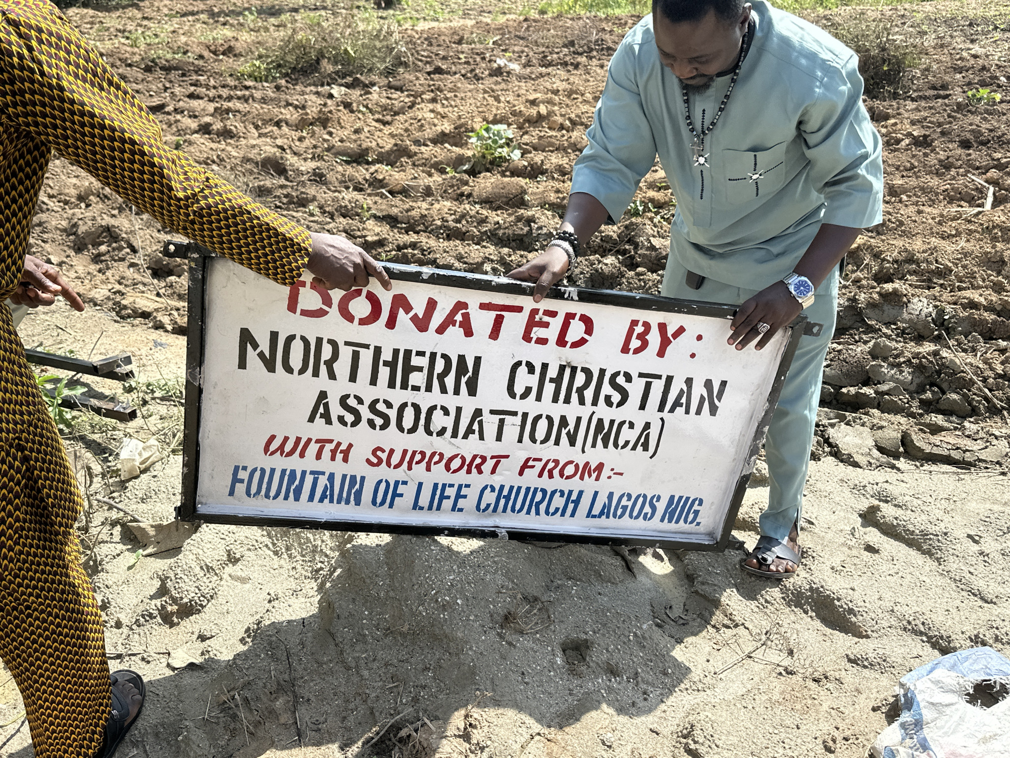 In Afogo, Nigeria, a white sign with red and blue lettering reads "DONATED BY: NORTHERN CHRISTIAN ASSOCIATION (NCA) WITH SUPPORT FROM: FOUNTAIN OF LIFE CHURCH LAGOS NIG." Two individuals are present. One, wearing a yellow and brown patterned garment, points to the sign. The other, in a light blue outfit and wearing jewelry and a watch, holds the sign. They are in an outdoor, dusty environment with tilled earth and sparse vegetation.