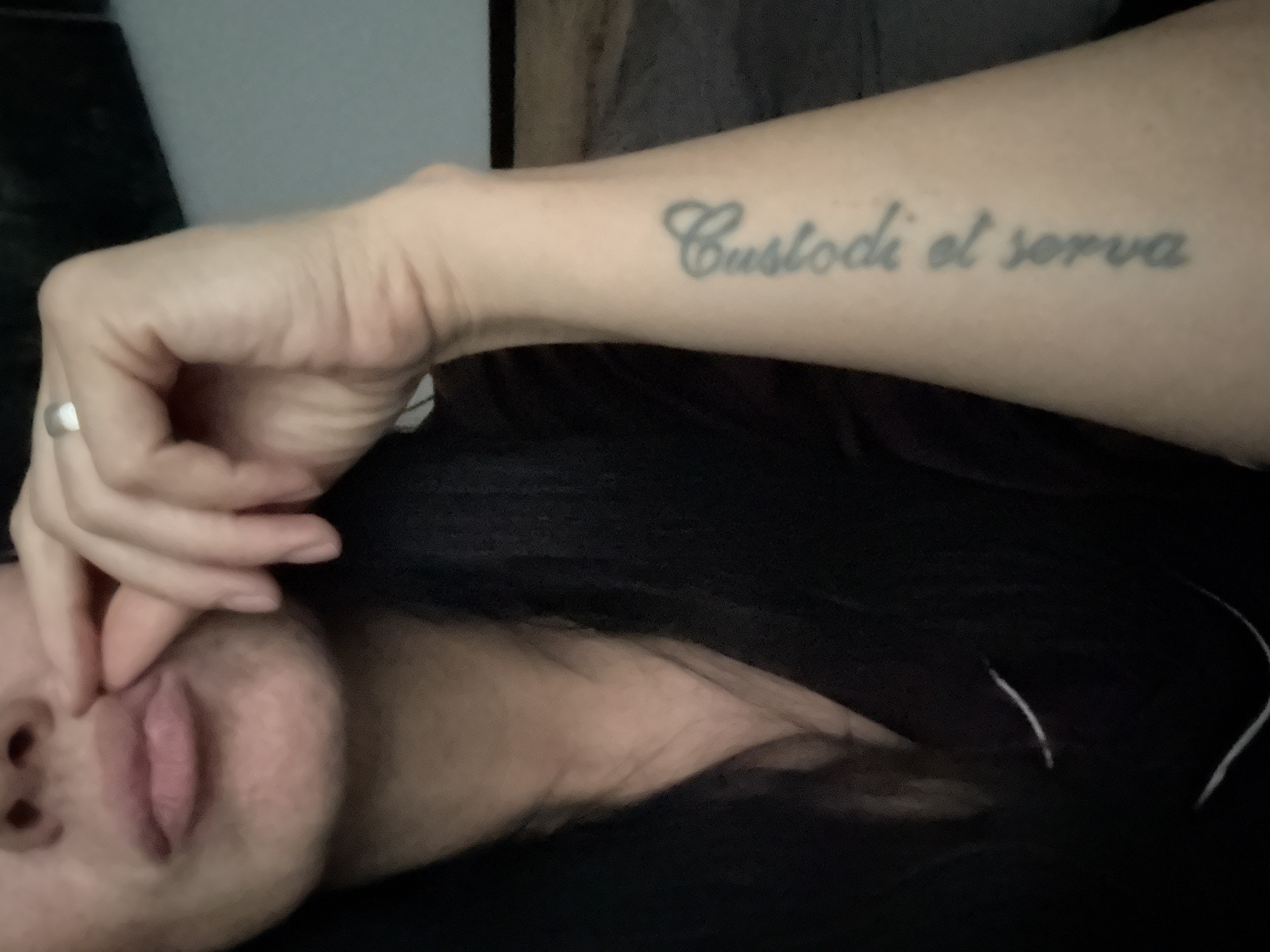 The image shows a close-up of a person's arm with a tattoo and their mouth and chin. The tattoo is on the person's forearm and reads "Cassiodi el sopra" in cursive script. The person's hand is near their mouth, and they are wearing a ring on their index finger. The background is dark, suggesting an indoor setting. There is no discernible information in the image that would allow for the identification of the location in the city of Chaikovsky, Russia.