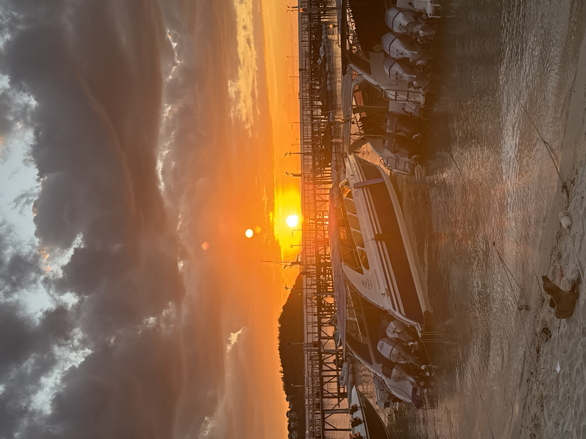 This image captures a vibrant sunset over Baan Bang Rak, Thailand. The sky is ablaze with shades of orange and yellow, illuminated by the setting sun, which appears as two distinct orbs due to atmospheric conditions. Dark, dramatic clouds drift across the sky, adding depth and contrast to the colorful display.
In the foreground, several speedboats are docked in the shallow, reflective water. These boats are equipped with multiple outboard motors, indicating their capacity for speed and transport. Their sleek designs and white and blue hulls are partially illuminated by the warm sunset light, casting shimmering reflections on the water's surface.
A wooden pier stretches out into the water, lined with lights and structures, suggesting it's a busy hub for boat activity. People can be seen walking along the pier, silhouetted against the bright sky. On the shore, a sandy beach meets the water, with a brown dog lying on the sand, looking towards the water. Further back, a green, hilly landscape with buildings can be seen in the distance, hinting at the coastal town. The overall scene evokes a peaceful yet active coastal evening.