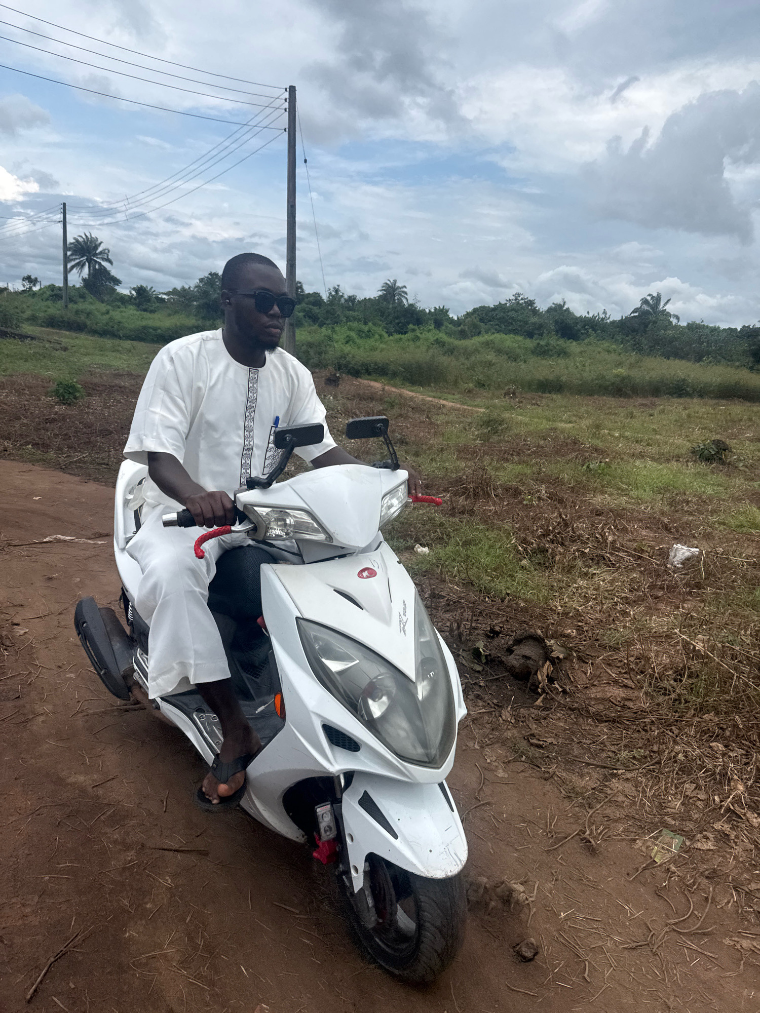 In Ihiagwa, Nigeria, a Black man wearing sunglasses and a white traditional outfit is riding a white scooter on a dirt road. The scooter has a red grip on the handlebar and a blue pen clipped to the rider's shirt. The background consists of a grassy field with scattered dry vegetation, palm trees, and dense foliage. Overhead, a cloudy sky with patches of blue is visible, with power lines and utility poles also present. The road appears muddy in places.