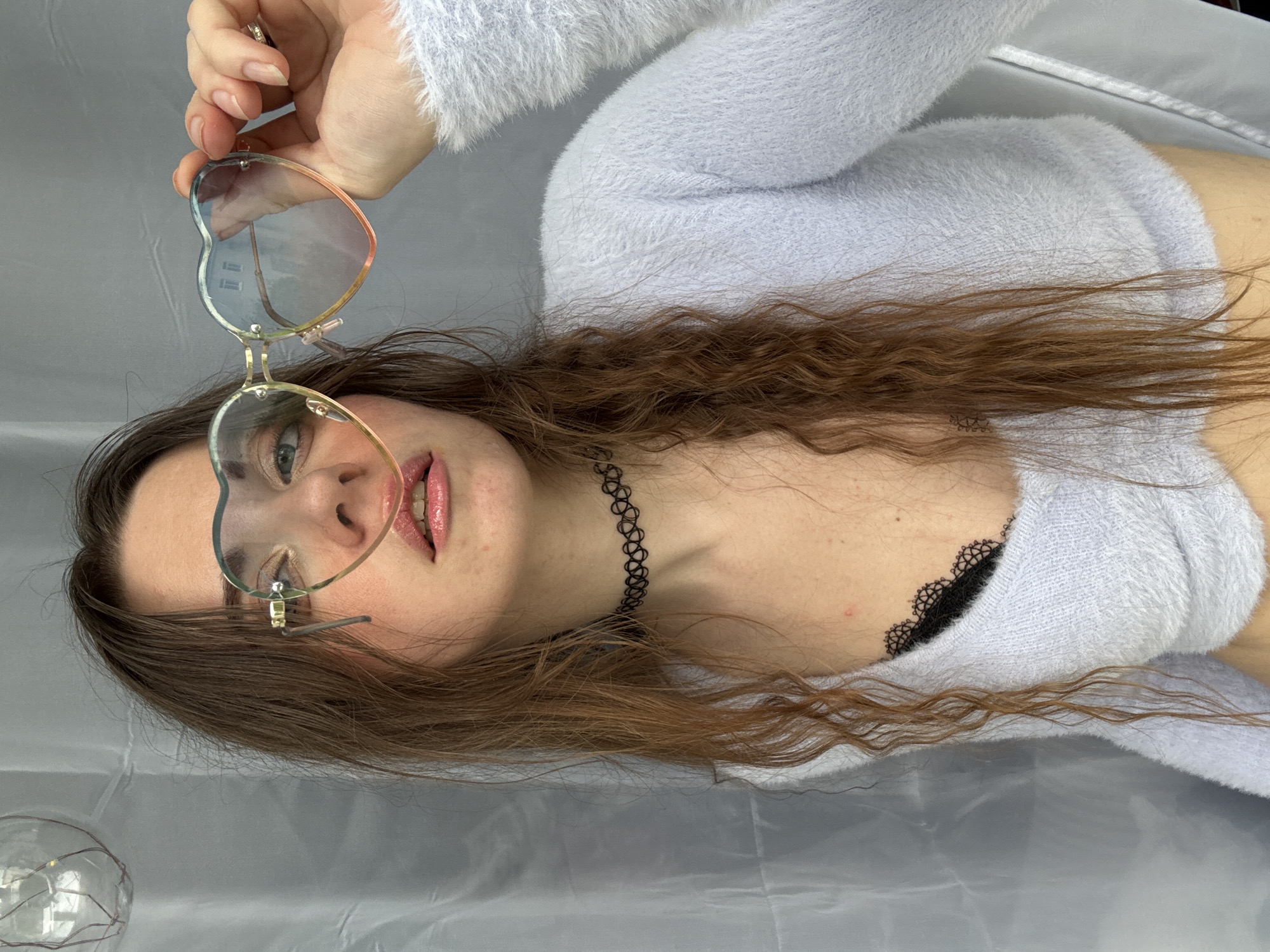 A young woman with long, wavy brown hair lies on a light gray surface, possibly a bed or a backdrop. She is wearing a light blue, fuzzy sweater and a black choker necklace. Her eyes are green, and she is wearing heart-shaped glasses with clear lenses and a colorful rim. Her lips are parted, and she is looking at the camera. A bit of black lace, possibly from a bra, is visible peeking out from under her sweater. In the bottom left corner, a clear glass sphere with some wire inside is partially visible. The lighting suggests an indoor setting, likely with soft, diffused light. No specific time of day or weather conditions are indicated. No visible text is present in the image. The overall mood is relaxed and playful.