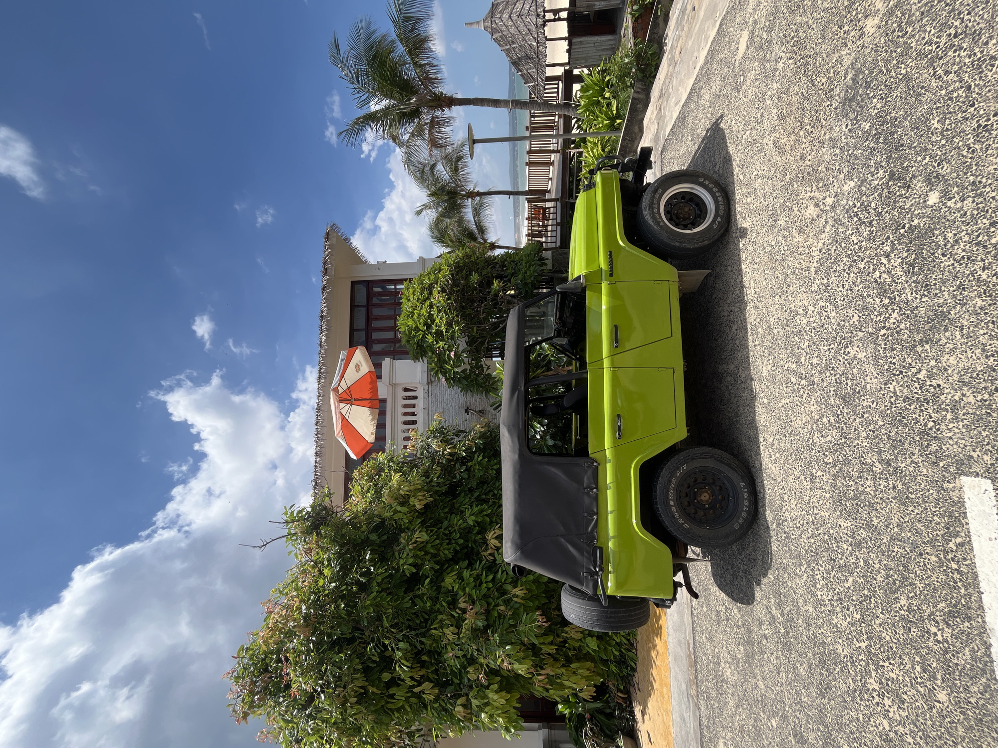 A bright green, open-top jeep is parked on a textured, asphalt surface. The jeep has a black fabric top that is partially lowered. The vehicle is positioned diagonally in the frame, with its front facing towards the upper left. In the background, there's a building with a thatched roof and white balconies, surrounded by lush green foliage and palm trees. An orange and white umbrella is visible near the building. Beyond the trees and building, a glimpse of the ocean and a wooden railing suggest a coastal setting. The sky is a vibrant blue with scattered white clouds, indicating a sunny day. The overall scene evokes a tropical, vacation-like atmosphere. The time of day appears to be midday or early afternoon, given the bright sunlight and the shadows cast by the jeep and trees. There is no visible text in the image. The location context provided is Phu Thuy Ward, Vietnam.