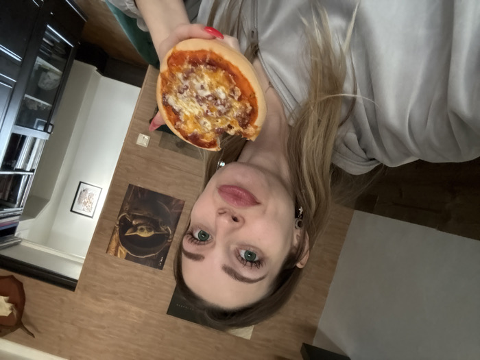 A fair-skinned woman, estimated to be in her 20s or 30s, with long blonde hair and green eyes, is depicted in a selfie taken indoors in Reutov, Russia. She wears a light grey zippered hoodie and black earrings. Her lips are slightly pouted as she directly faces the camera. In her right hand, adorned with red nail polish, she holds a partially consumed small round pizza topped with cheese and sauce. The background features a light brown wooden paneled wall with a poster of Grogu (Baby Yoda) and a white light switch. To the right, a darker door frame is visible, above which an animal skull or horn is mounted on a plaque. Further right, a dark, glass-fronted display cabinet or bookcase containing various items is present, with a small framed picture hanging on the wall beyond it.