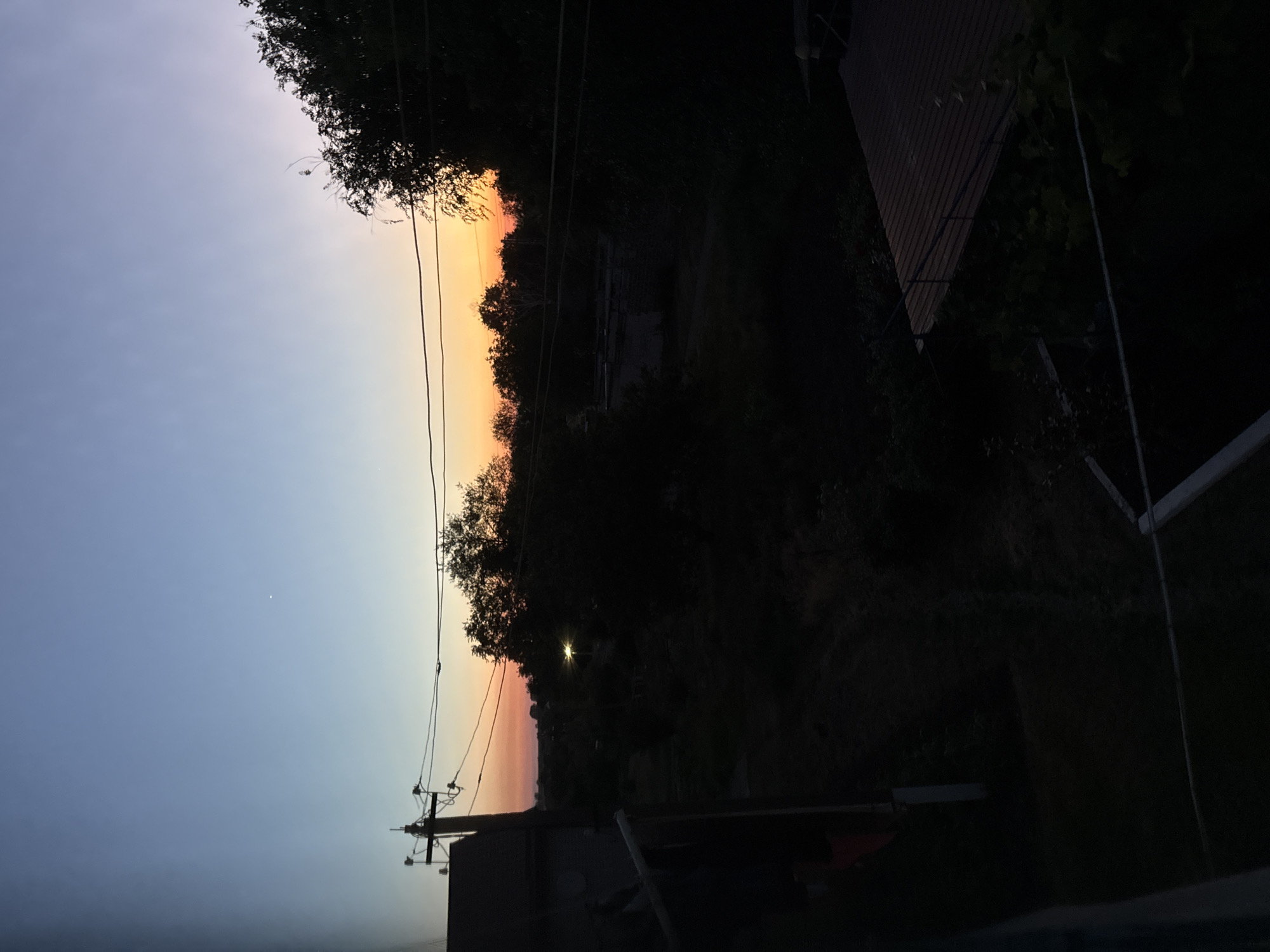 The image shows a view of a rural landscape at dusk, likely in a village or suburban area. The sky is transitioning from a deep blue to a soft orange and yellow near the horizon, indicating sunset or sunrise. Silhouetted trees and vegetation dominate the mid-ground and foreground, obscuring much of the detail of the landscape. A power line runs across the frame, supported by a utility pole with insulators. On the left, a dark, angular structure, possibly a building or shed, is partially visible. In the distance, a solitary light bulb illuminates a small area, casting a faint glow. The overall impression is one of twilight and quietude.