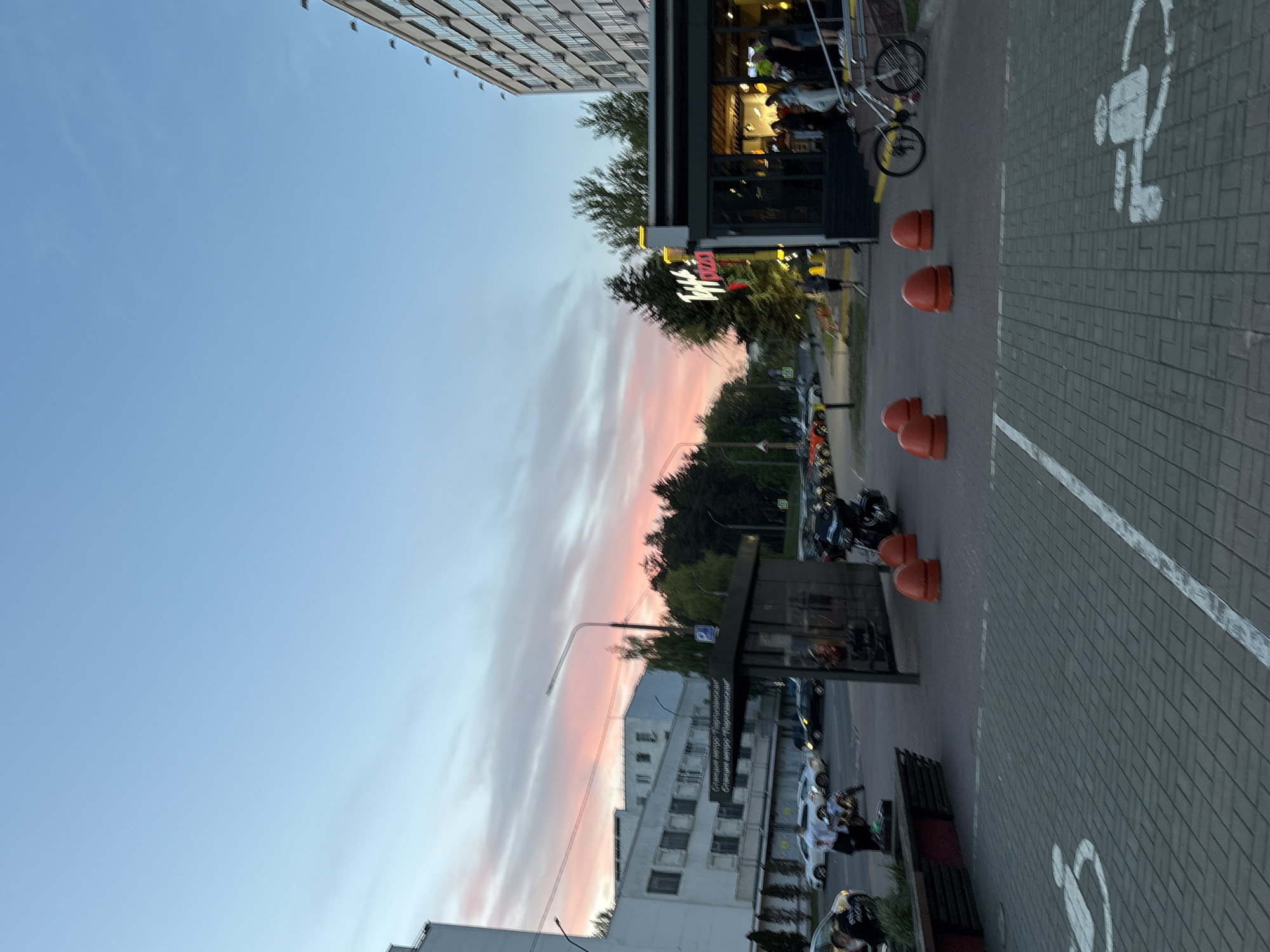 The image shows a street scene in Minsk, Belarus, at dusk. The sky is a mix of blue and pink hues, suggesting either sunrise or sunset. A tall building with multiple windows is visible on the left side of the frame. On the right side of the frame, there's a restaurant with a sign written in Cyrillic script above its entrance. An orange sign is also visible. Parking cones and a row of parked motorcycles are present along the street. The street surface is paved with gray bricks, and there are white markings indicating parking spaces for people with disabilities. Further down the street, more buildings are visible, along with trees. People are seated outside of the restaurant.