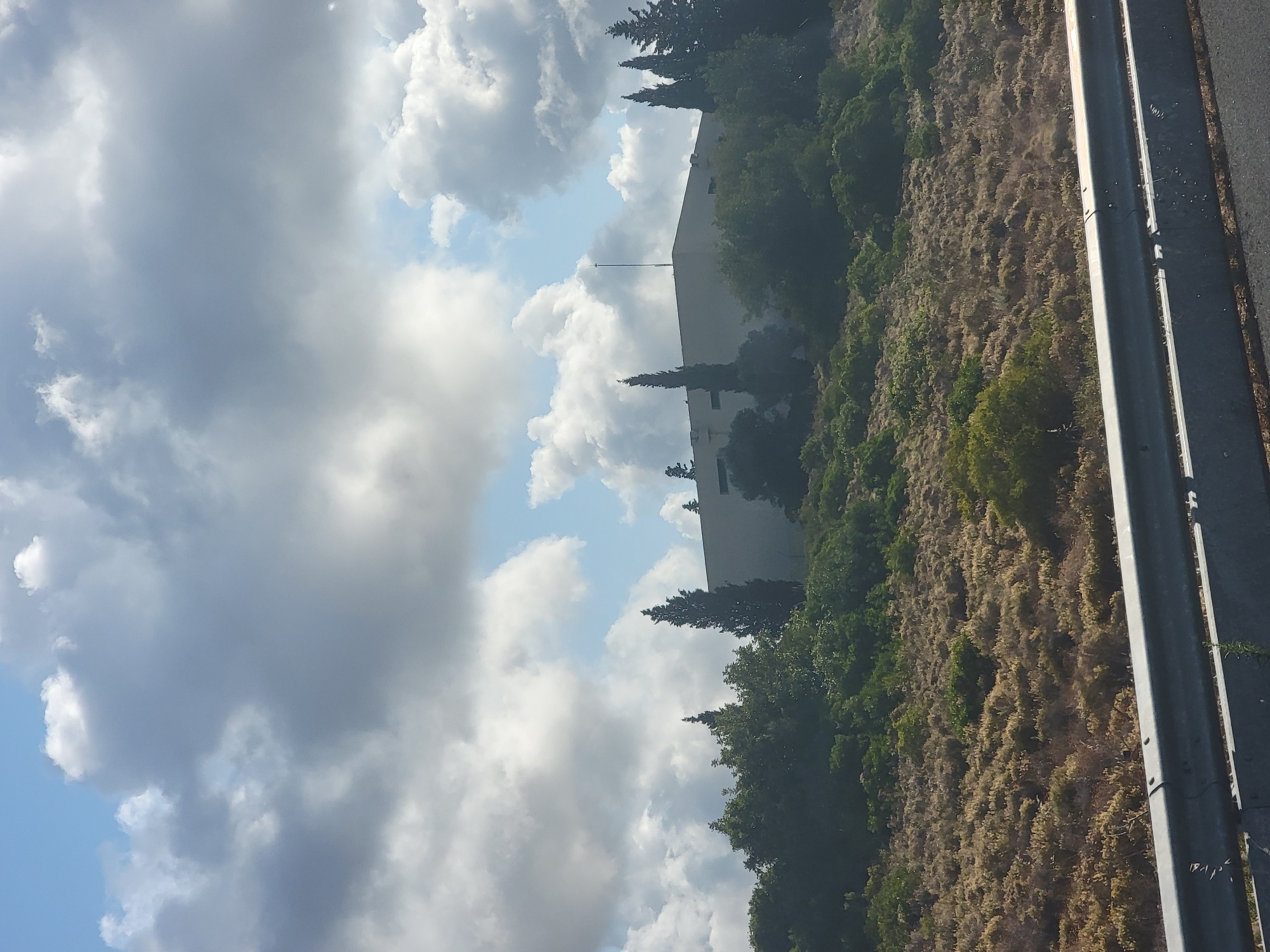 The image depicts a building situated on a hill, partially obscured by trees and vegetation. The building is a light-colored, rectangular structure with a flat roof and a single antenna on top. The surrounding landscape is covered with dry, brush-like vegetation and scattered green trees, creating a natural, somewhat arid environment. In the foreground, a metal guardrail is visible, indicating the presence of a road. The sky is partly cloudy, with a mix of white and gray clouds against a blue sky. The overall impression is of a suburban or rural setting, possibly on the outskirts of Almancil, Portugal, given the hint provided, where the natural landscape meets a man-made structure. No people are visible in the image, and no specific actions or interactions are depicted.