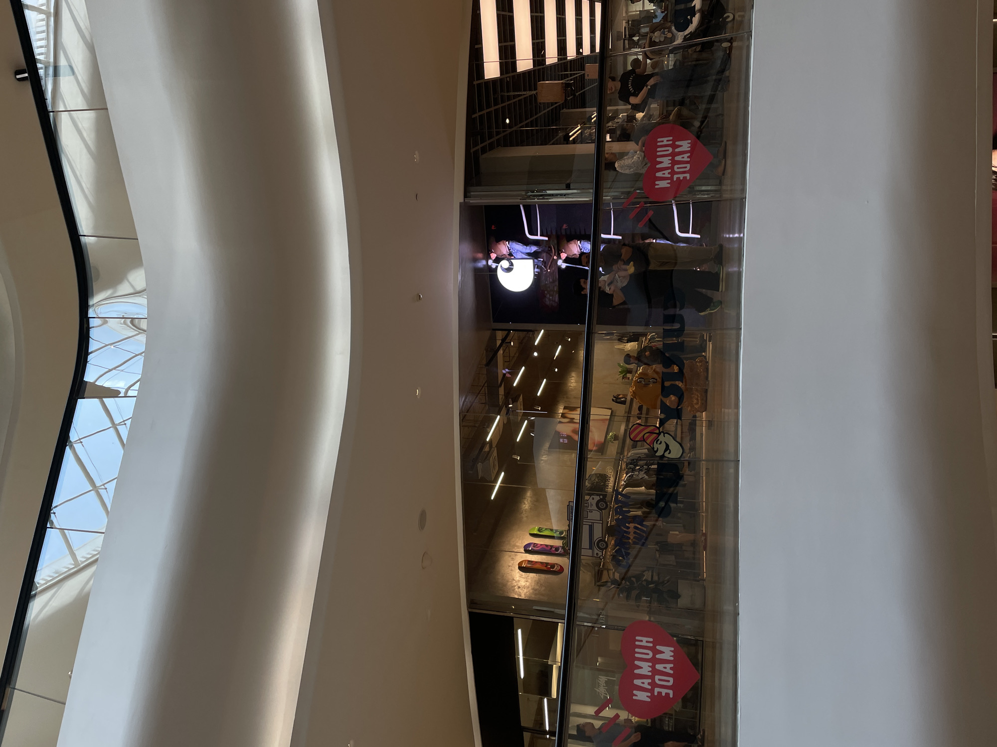 The image captures a modern, multi-level indoor space, likely a shopping mall or a large retail establishment, within Bangkok, Thailand. The dominant features are large, smoothly curved, light-colored architectural elements on the left, contrasting with a clear glass facade on the right that provides a view into an enclosed area or store.
Inside the glass-encased area, several individuals are visible, casually dressed, appearing to be browsing or observing. The space features modern design with a staircase and prominent strip lighting on the ceiling, creating bright reflections on the glass. A distinctive large, circular light fixture is also visible. Merchandise, including several colorful skateboards mounted on a wall, can be seen on display.
Two notable red heart-shaped signs are positioned inside the store, reflecting off the glass. The text on these signs, when correctly oriented and read from the inside out, spells "HUMAN MADE," but appears as a reversed "WAMUH 3DAM" due to reflection. This brand suggests a fashion or lifestyle retail space.
In the background, visible through a window-like opening on the far left, is a complex, geometrically patterned skylight, allowing natural light to enter the building and suggesting it is daytime. The overall ambiance is bright and contemporary, characteristic of a high-end commercial environment.