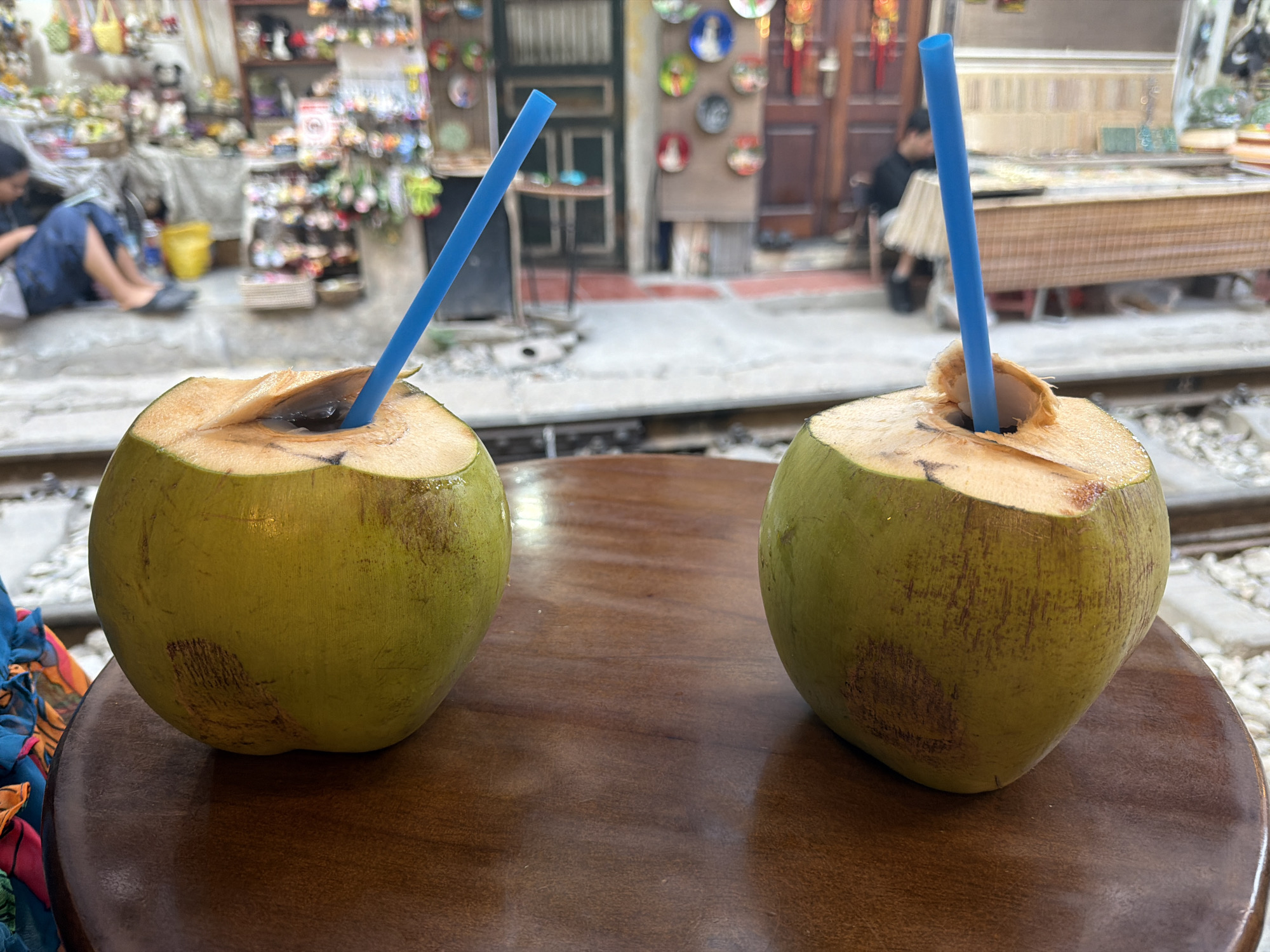 Two green coconuts with blue straws are placed on a round wooden table. The coconuts appear to have been cut open for drinking. Behind the coconuts, the scene includes a blurry backdrop of a city street with shops and pedestrians. The setting is likely the "Train Street" in Hà Nội, Vietnam, as evidenced by the presence of train tracks in the immediate background. People are seated or walking along the sides of the tracks amidst shops and residential buildings. Decorations and wares for sale are visible in the shops, suggesting a tourist-oriented area.