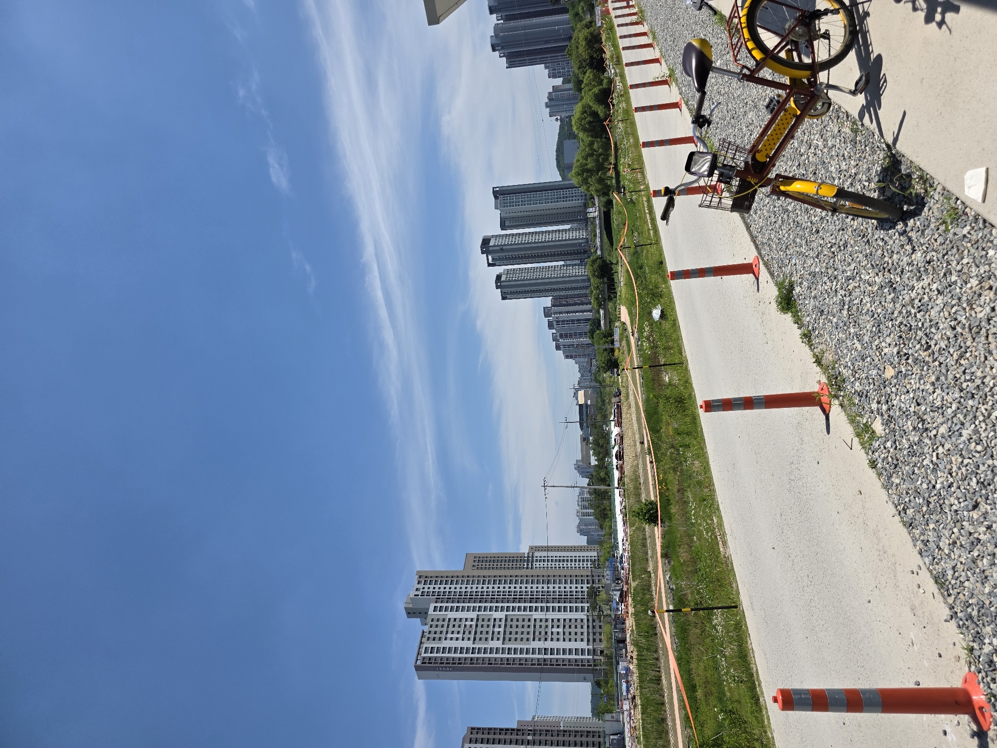 The image depicts an outdoor scene in a developing urban area, likely in Daejeon, South Korea, given the architectural style of the buildings. In the foreground, a bright yellow and brown bicycle is parked on a gravel path beside a paved walkway. The bicycle has a basket on the front and a rear rack. Orange traffic cones with reflective gray bands are spaced along the edge of the walkway, demarcating a construction or safety zone.
Beyond the walkway, there's a grassy area with some vegetation and orange construction fencing. In the mid-ground, several tall, modern apartment buildings are visible, some with distinctive gray and white facades. Further in the distance, more city buildings and some green trees are scattered across the landscape. The sky is a clear, bright blue with a few wispy white clouds. The overall impression is a sunny day in a city undergoing construction and development.