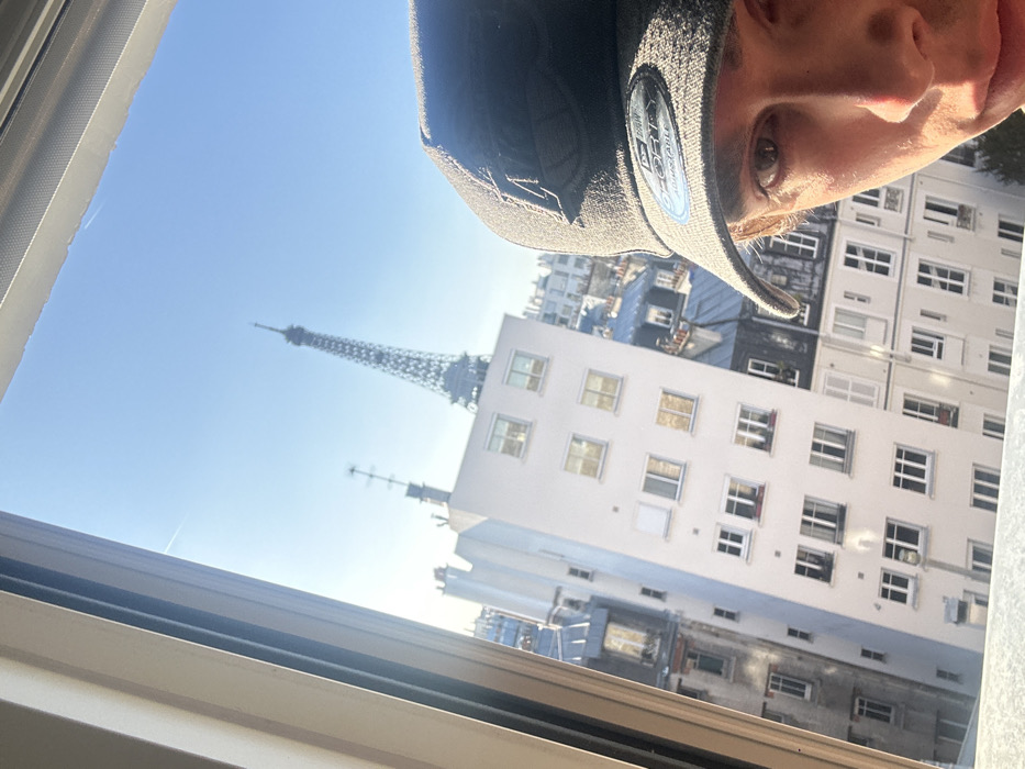The image depicts a partial selfie taken from an elevated interior position, looking out a window onto a Parisian cityscape. In the bottom right quadrant, the right side of an individual's face, specifically the right eye, cheek, and jawline, is visible. The individual is wearing a dark baseball cap with a dark stylized logo on the front and a "9 FORTY ADJUSTABLE" sticker on the brim, partially shadowing their face. A light-colored window frame runs vertically along the left edge and horizontally across the top of the image.
Outside the window, a bright, clear blue sky occupies the upper background. The Eiffel Tower is prominently centered in the background, its latticework structure distinctly visible against the sky. Below the Eiffel Tower and extending to the left, a multi-story, light-colored building with numerous rectangular windows is present. Beyond this, classic Parisian rooftops with dark, sloped surfaces, chimneys, and attic windows are discernible. An antenna structure stands atop a building to the left of the Eiffel Tower. The scene is illuminated by bright daylight, suggesting clear weather conditions. The identifiable landmark confirms the location as Paris, France.