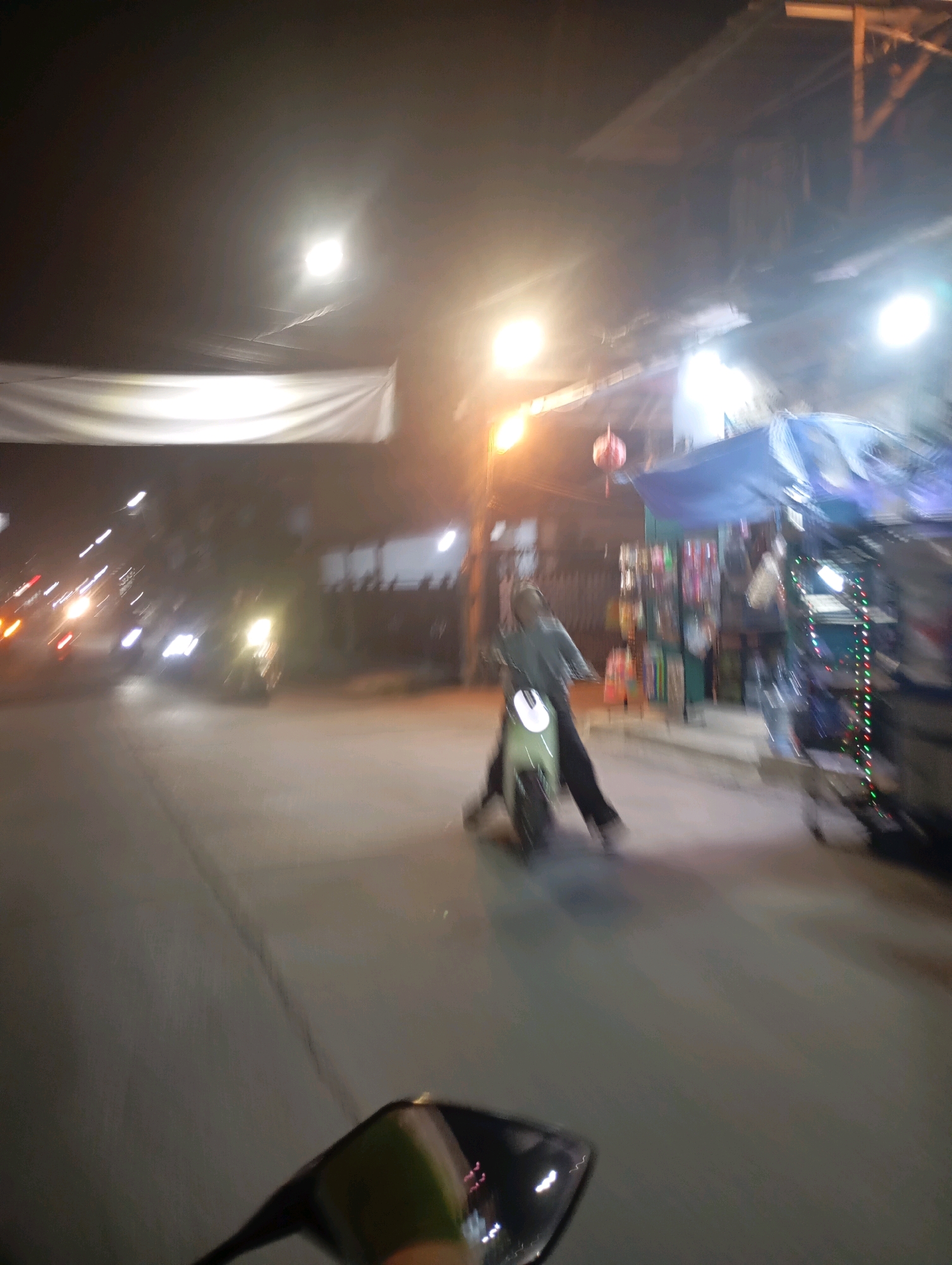 This image captures a blurry, night-time scene on a street in Babelan, Indonesia. In the center, a person is riding a light-green scooter. The rider is wearing dark pants and a light-colored, possibly gray, hooded jacket. The scooter has a visible headlight. To the right of the scooter, there appears to be a roadside stall or cart decorated with colorful lights. The background shows streetlights, buildings, and a white banner stretched across what looks like a storefront. The road is paved, and there are other blurred lights in the distance, suggesting traffic. The overall impression is that of a busy urban environment at night.