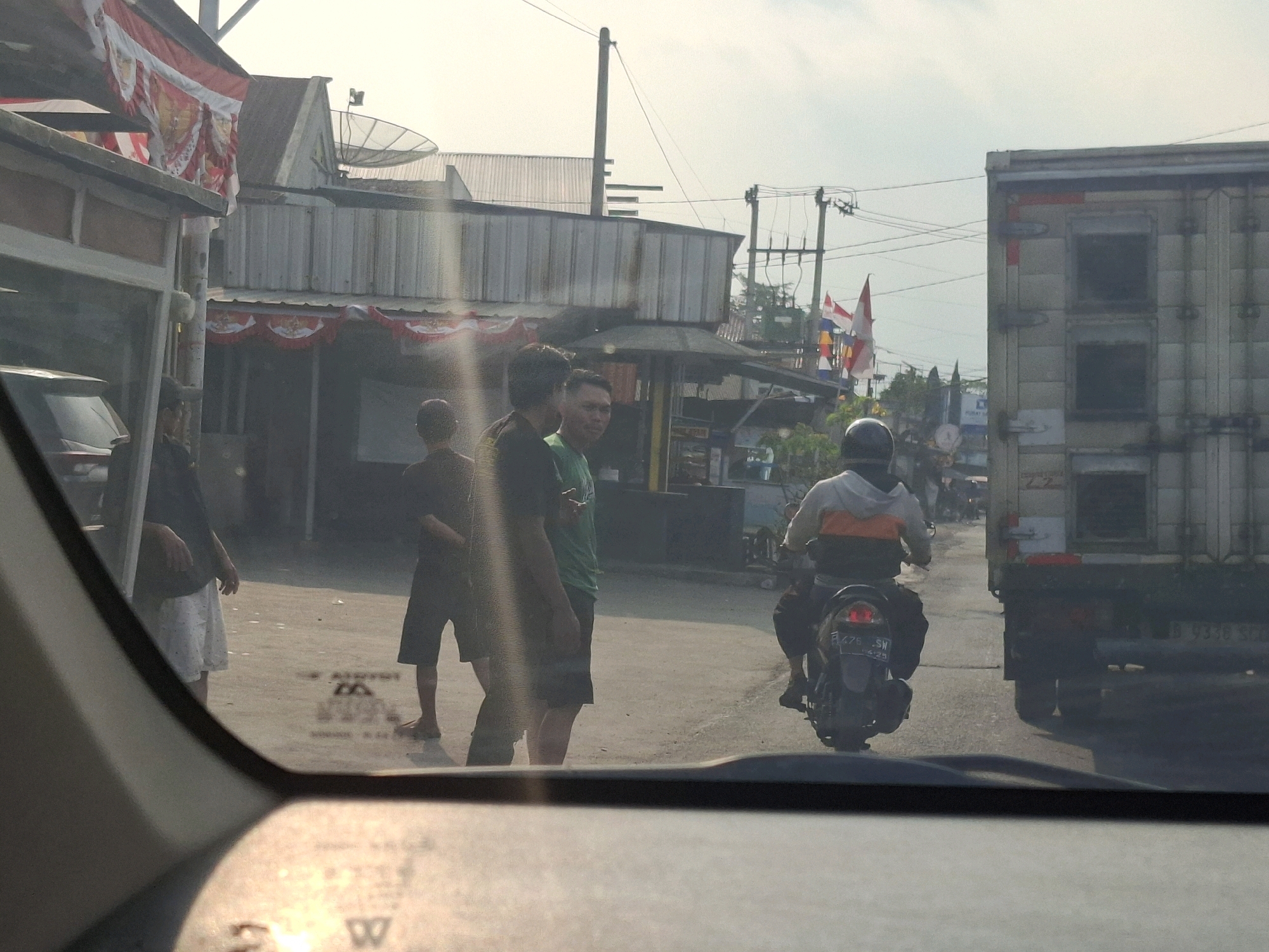 The image depicts a street scene in Sukabumi, Indonesia, as seen through a car windshield. The time appears to be daytime, with bright sunlight casting lens flares across the view. In the foreground, the car's dashboard and a portion of the windshield are visible, including a reflection of what might be a street sign.
In the mid-ground, several individuals are present. Three men are standing on the roadside, one in a dark shirt and white shorts, another in a dark shirt and dark shorts, and a third in a green t-shirt and dark shorts. To the right, a person is riding a motorcycle, wearing a hooded jacket with orange and white accents, and dark pants. They are traveling in the same direction as the vehicle from which the photo is taken.
Behind the people, there are buildings with corrugated metal roofs and various awnings. Indonesian flags, red and white, are displayed, some as bunting and others as larger flags on poles. A large truck, likely a delivery or cargo truck, is to the right of the motorcycle, also traveling in the same direction. The truck is partially obscured by the motorcycle and the perspective of the shot. Electrical poles and wires are visible in the background, suggesting an urban or semi-urban environment. The road surface is asphalt, with some visible markings. The overall atmosphere suggests a typical street scene in a developed area of Indonesia, possibly a commercial or residential district.