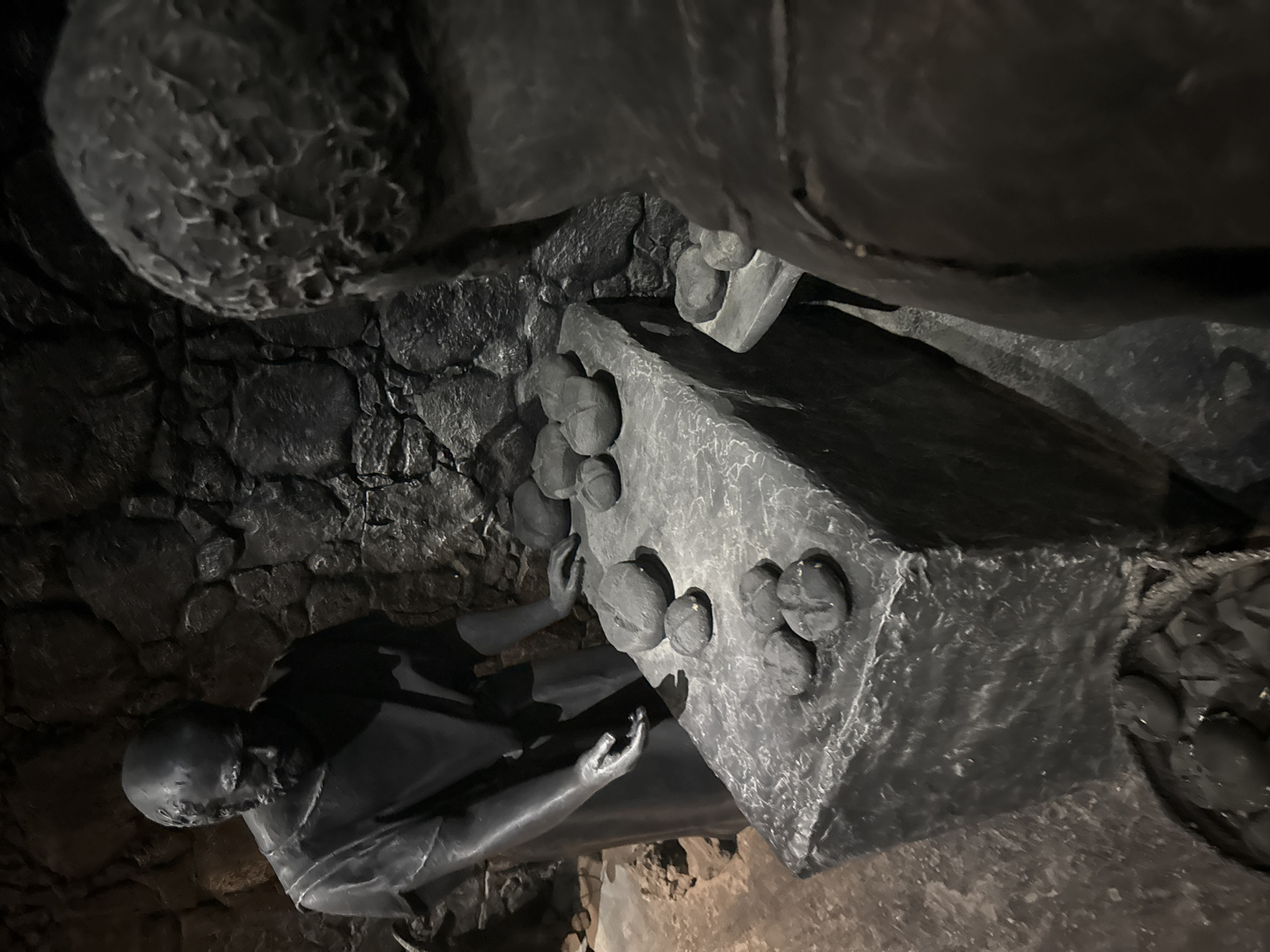 In the city of Tamar Regional Council, Israel, a dark, cave-like environment features a diorama of two figures in dim lighting. The scene depicts two bronze-colored statues, possibly representing bakers or historical figures. One statue, on the left, is a man with a beard, wearing a draped garment, standing beside a stone table. He is looking down at the table, with one hand outstretched, as if to touch or place bread. The stone table is laden with multiple round loaves of bread, also rendered in bronze. Another statue, partially visible in the foreground on the right, faces away from the viewer and towards the table, its back and shoulder in view. The background is composed of rough, uneven stone walls, reminiscent of a cave or ancient dwelling. A woven basket filled with more bronze loaves is visible at the base of the stone table. The overall impression is one of a historical or religious tableau, possibly illustrating an event involving bread-making or distribution.