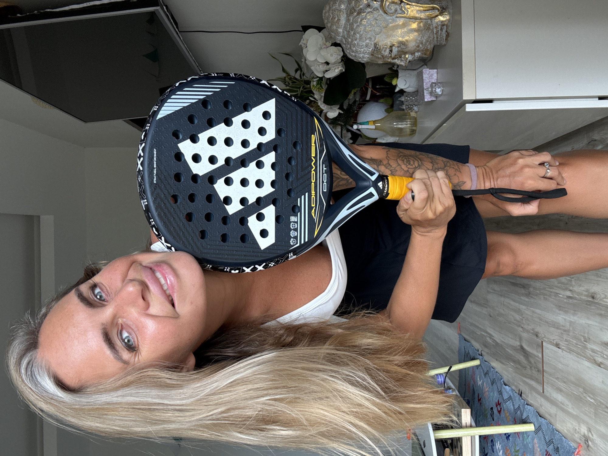 A Caucasian woman with long blonde hair, partially graying, and blue eyes is leaning forward, holding an Adidas padel racket. She is wearing a white tank top and a black skirt. Her left arm, tattooed with floral designs, is wrapped around the racket. She is smiling at the camera. The background includes a television, a cabinet with decorative items including a gold-colored Buddha head statue and white orchids, and a patterned rug on a wooden floor. The scene appears to be indoors, possibly in a residential setting. The location is not determinable from the image alone.
