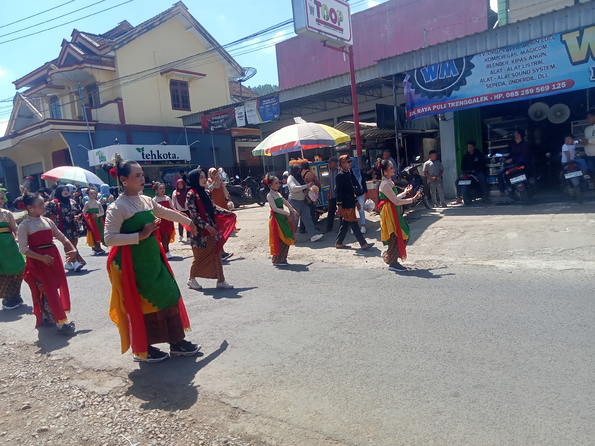 A street-level, eye-level medium shot captures a procession of young women in traditional Indonesian attire walking on a paved road. The women, adorned in colorful skirts and tops, move from left to right across the frame, their bodies angled slightly towards the viewer. Their attire consists of vibrant red, yellow, and green fabrics, some draped over their shoulders, creating a dynamic visual. They are performing in what appears to be a parade or cultural event.
The backdrop of the scene includes buildings with distinct architectural styles, suggesting an urban or semi-urban environment. On the left, a building with a yellow facade and a dark blue base stands prominently, featuring traditional Indonesian roof designs. Further down the street, a business with a large banner displaying "WM" and listing various electronic goods and services is visible. A prominent "TOOP" sign is mounted on a pole above the street.
Several other individuals, including men and women, are present on the sidewalks and along the street, observing the procession or engaged in other activities. Some are standing near parked motorcycles, while others are walking. The environment is sunny, with clear blue skies, indicating fair weather. The road surface is asphalt, with some gravel and debris on the side. The overall scene depicts a lively community event in Pule, Indonesia.