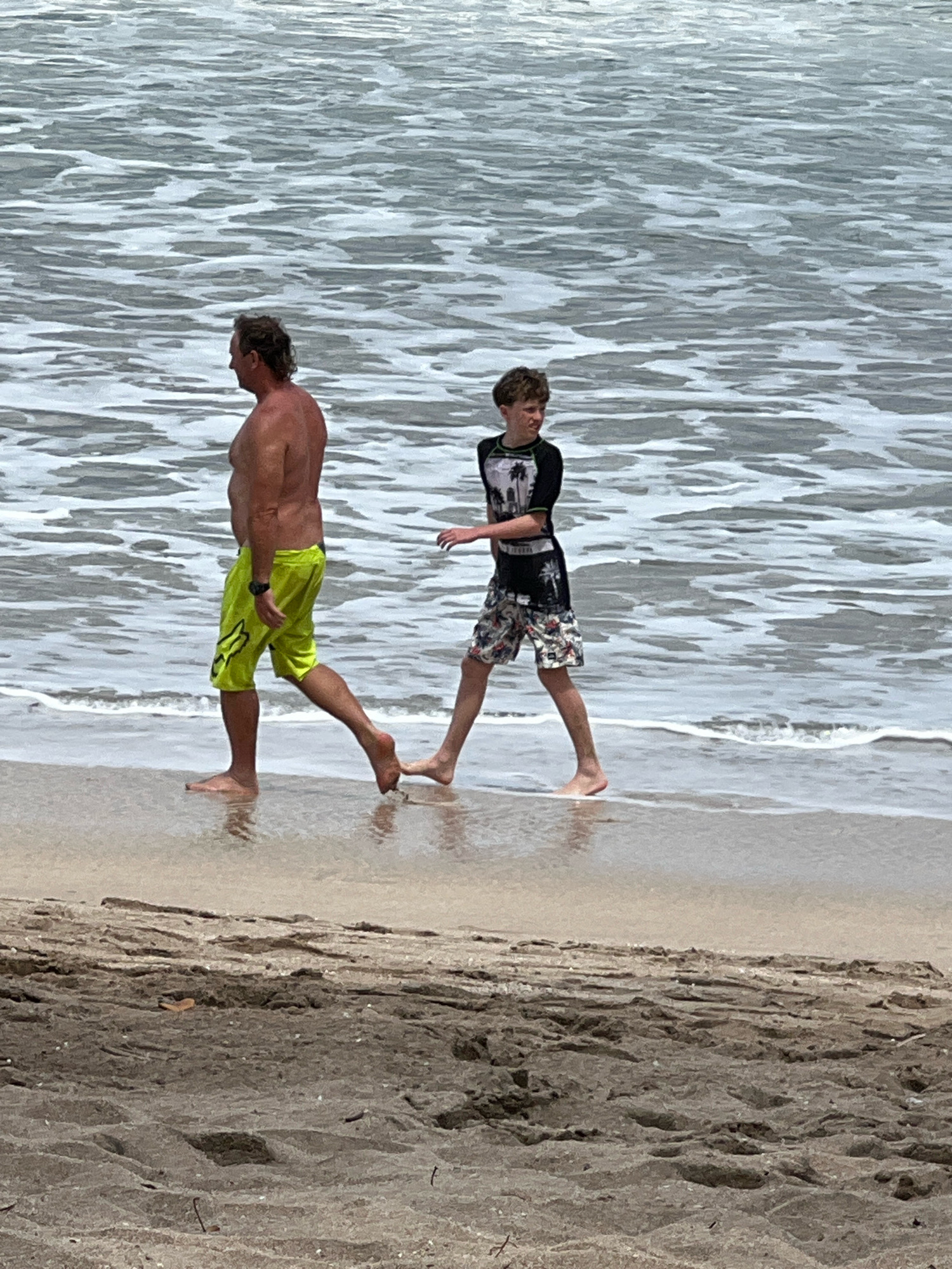 Two individuals, an adult male and a juvenile male, are walking on a sandy beach adjacent to the ocean. The adult male, shirtless and wearing lime green swim trunks, is walking to the left of the frame. The juvenile male, wearing a rash guard and patterned swim trunks, is walking to the right of the adult. The ocean water is visible in the background, with gentle waves reaching the shore. The foreground shows the sandy beach with some debris. The lighting suggests an overcast or cloudy day. This scene is consistent with the Kuta Beach area in Kuta, Indonesia.
