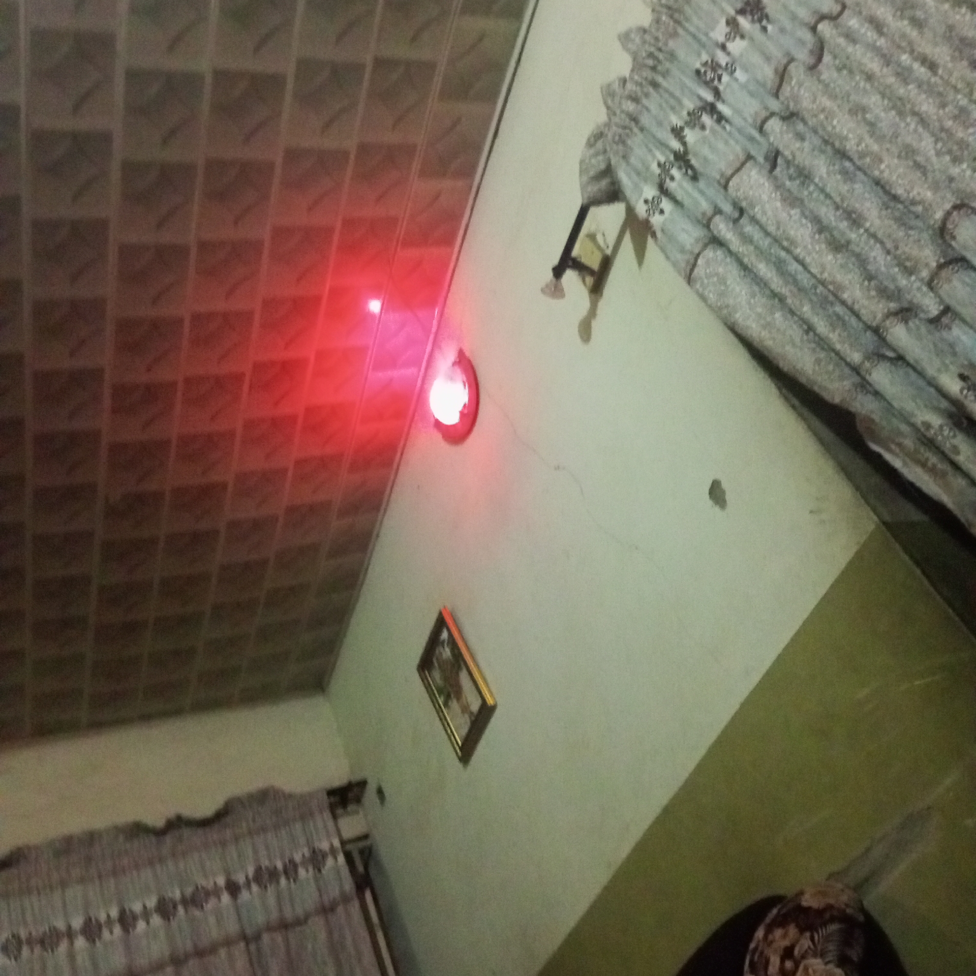 This image depicts an indoor scene in Howayi, Nigeria. The room appears to be dimly lit, with a prominent red light emanating from a fixture on the wall, likely an alarm or indicator light. A ceiling with a tiled pattern is visible in the upper left portion of the frame, also illuminated by the red light. To the right, a light-colored wall features a framed picture hanging on it. Further to the right, a patterned curtain is drawn, suggesting windows are present. The lower part of the image shows more of the patterned curtain and a portion of the floor or a surface covered by dark material. The overall atmosphere is somewhat obscured by the lighting and angle, but it suggests an interior space within a residential or commercial building. No people are clearly visible, and the primary focus is on the ambient lighting and the decorative elements of the room.