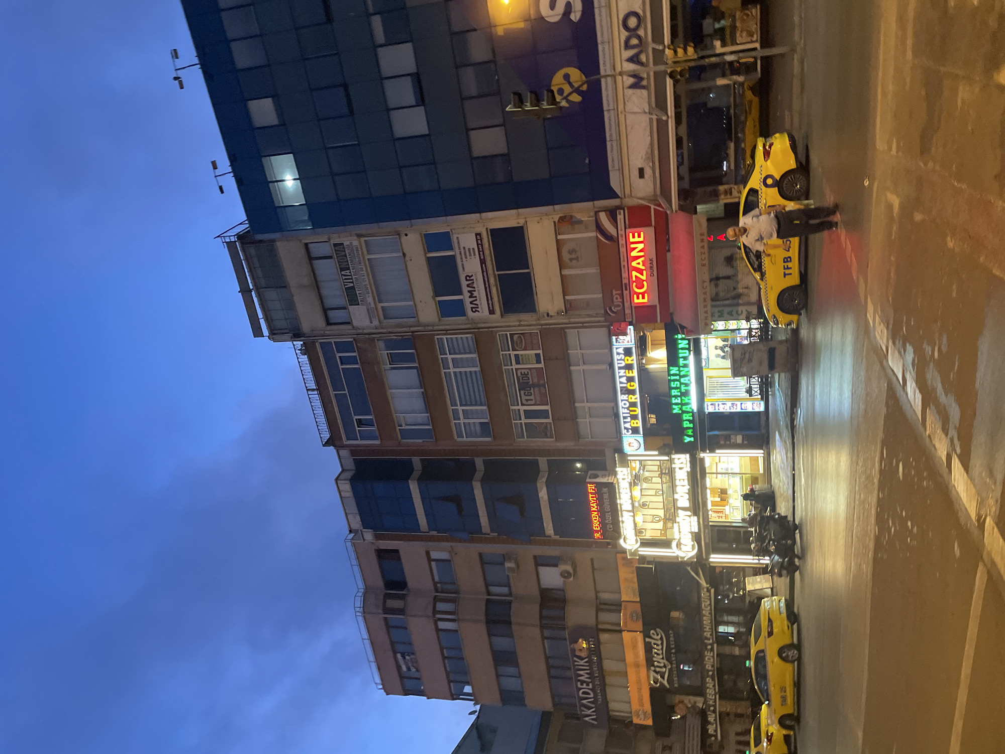 The image depicts a street scene in Kadıköy, Turkey, at dusk. Several multi-story buildings line the street, featuring a mix of commercial establishments on the ground floor and offices or apartments above. Prominent signage includes "AKADEMIK" (likely an educational institution), "Ziyade" (restaurant and kebab), "Kadıköy Börekçisi" (a pastry shop), "CALIFORNIAN BURGER", and "ECZANE" (pharmacy). Yellow taxis are visible, one of which is parked near a man standing in the street. Another taxi has the license plate "TFB 45". A motorcycle is parked on the sidewalk. The sky is a deep blue with wisps of clouds, indicating the transition from day to night. Streetlights and illuminated signs cast a warm glow on the wet pavement, suggesting recent rain. Traffic lights are visible. The architecture appears to be a mix of modern and older styles.
